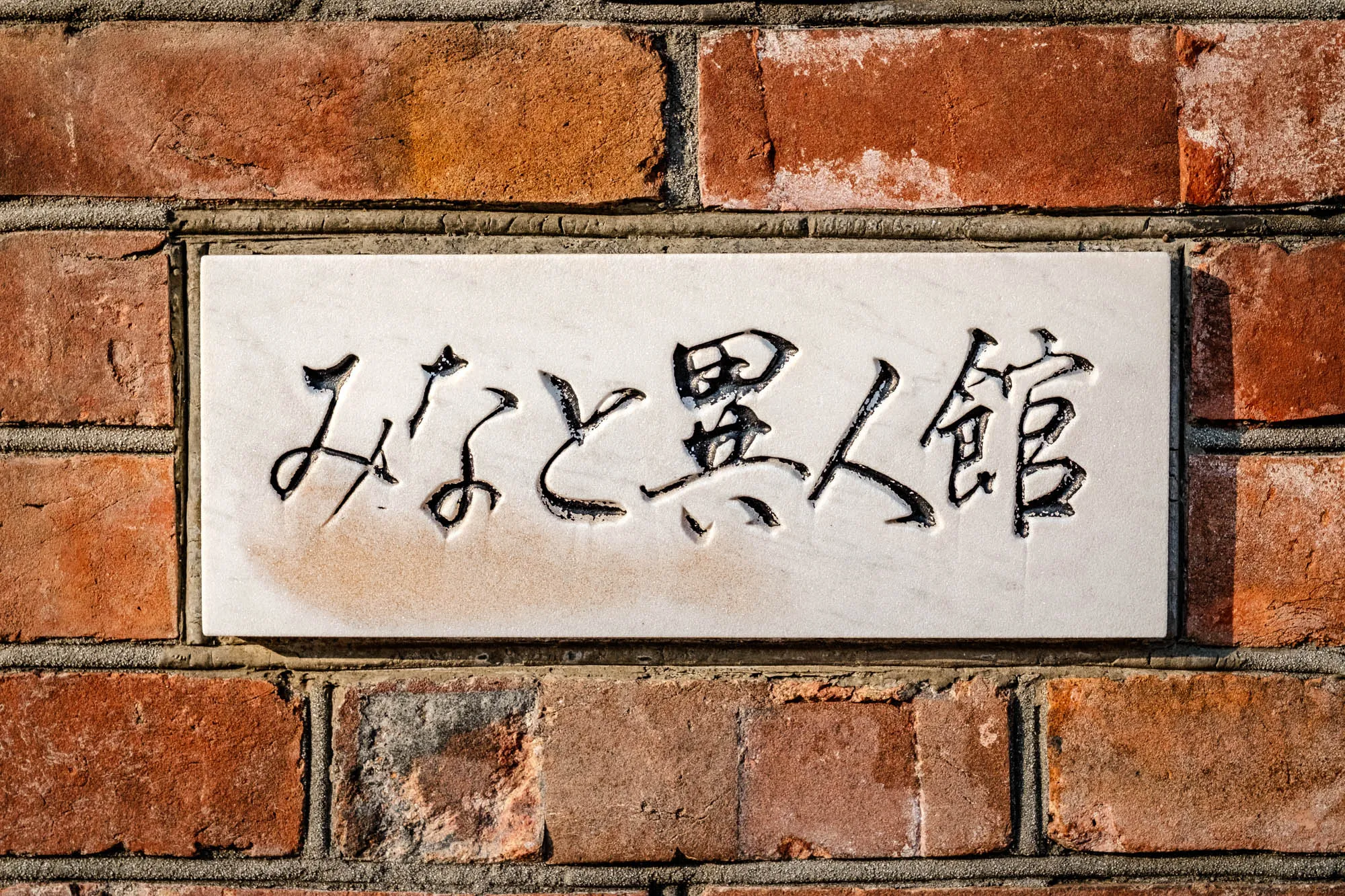 A rectangular plaque of smooth white stone is recessed into a wall of rough, weathered red brick. The Japanese inscription 'みなと異人館' is carved deeply into the center and filled with black ink, identifying the structure as the Minato Ijinkan. Sunlight from the upper right creates defined shadows within the strokes of the characters and along the uneven mortar joints of the surrounding masonry. The texture of the bricks varies from porous and pitted to smooth and fired, contrasting with the pristine surface of the signage.