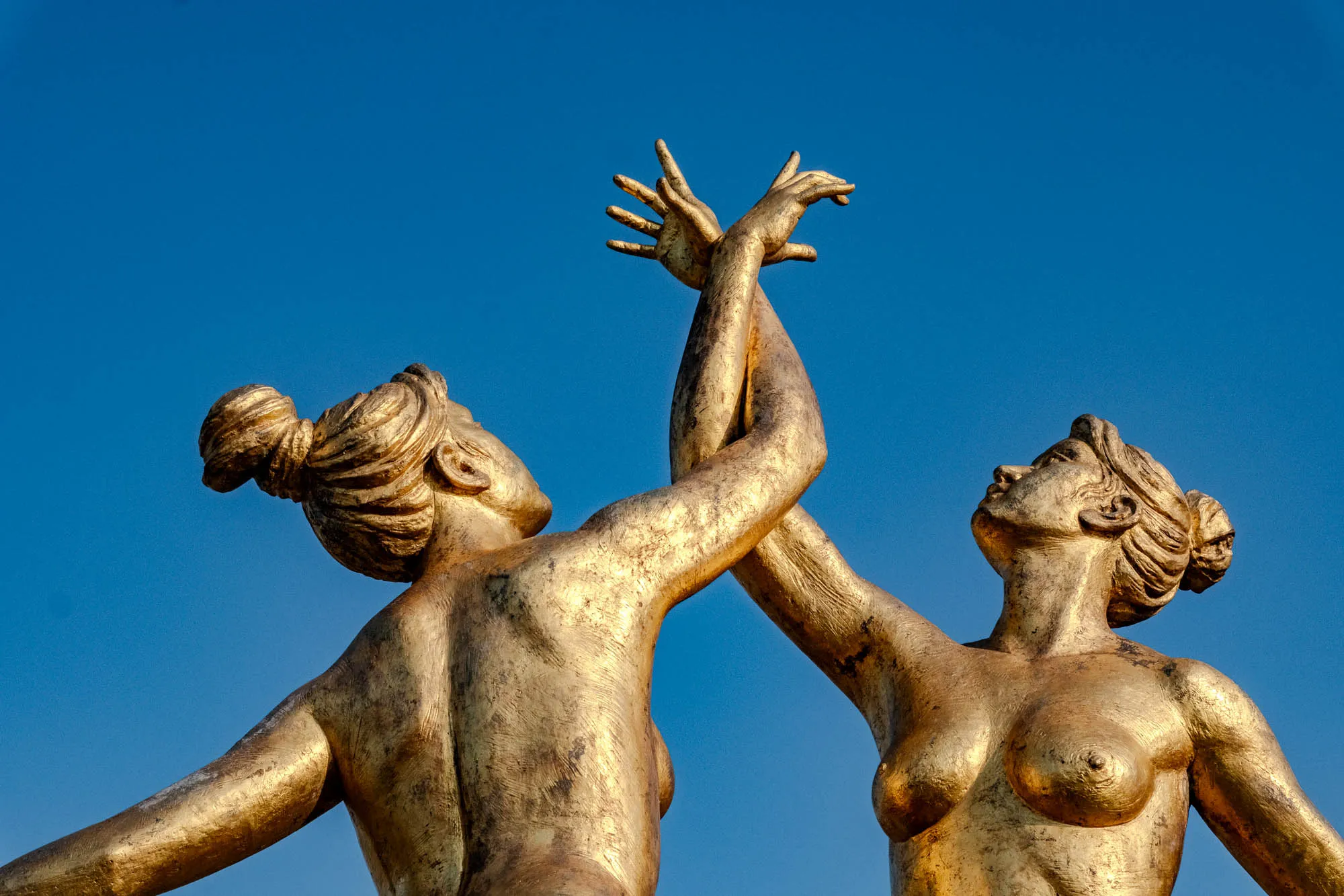 Two gilded female figures reach upward against a deeply saturated, cloudless blue sky, captured in a low-angle shot that emphasizes their verticality. The sculpture depicts one woman seen from behind and another facing forward, their arms raised and intertwined at the wrists in a gesture of connection. Harsh morning sunlight glints off the textured, metallic surface, highlighting the details of their braided hair and the anatomical curves of their torsos. Situated in the Kobe port district, the golden subject matter creates a complementary color contrast with the stark azure background.