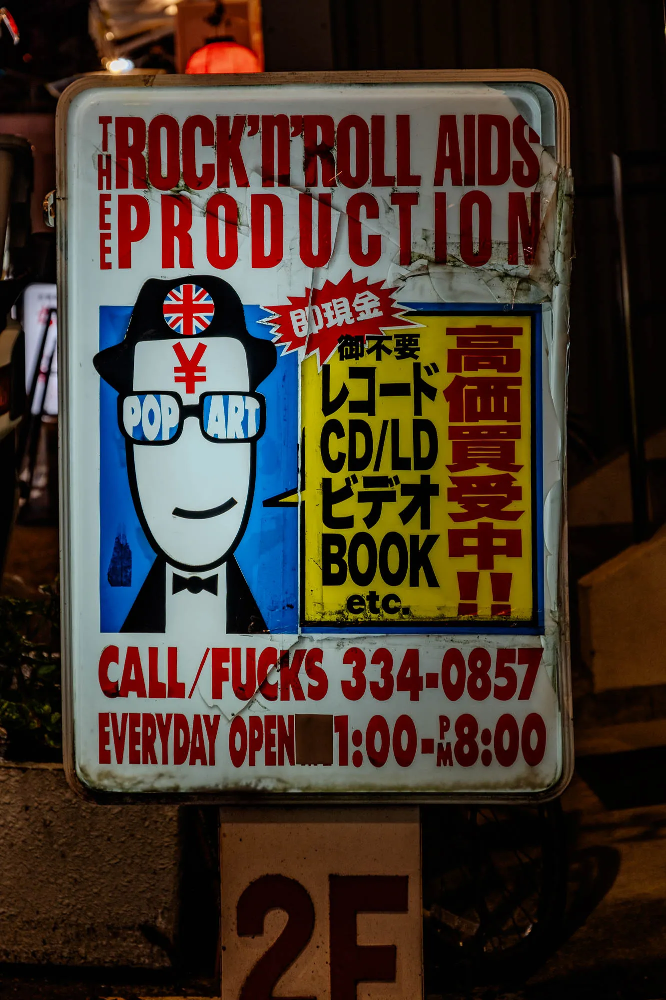 An illuminated, weathered street sign dominates a dark urban scene, featuring bold red and yellow text alongside a stylized cartoon figure. The white sign displays "THE ROCK'N'ROLL AIDS PRODUCTION" in distressed red letters at the top, while a blue panel on the left features a white-faced character in a black hat with a Union Jack badge, black sunglasses marked "POP ART," and a red Yen symbol on its forehead. To the right, a bright yellow section lists "CD/LD VIDEO BOOK etc." in Japanese and English, bordered by a red starburst graphic. The bottom of the sign includes red text reading "CALL/FUCKS 334-0857" and "EVERYDAY OPEN 1:00-PM 8:00," its edges peeling and grimy from age, with a "2F" marker visible below.