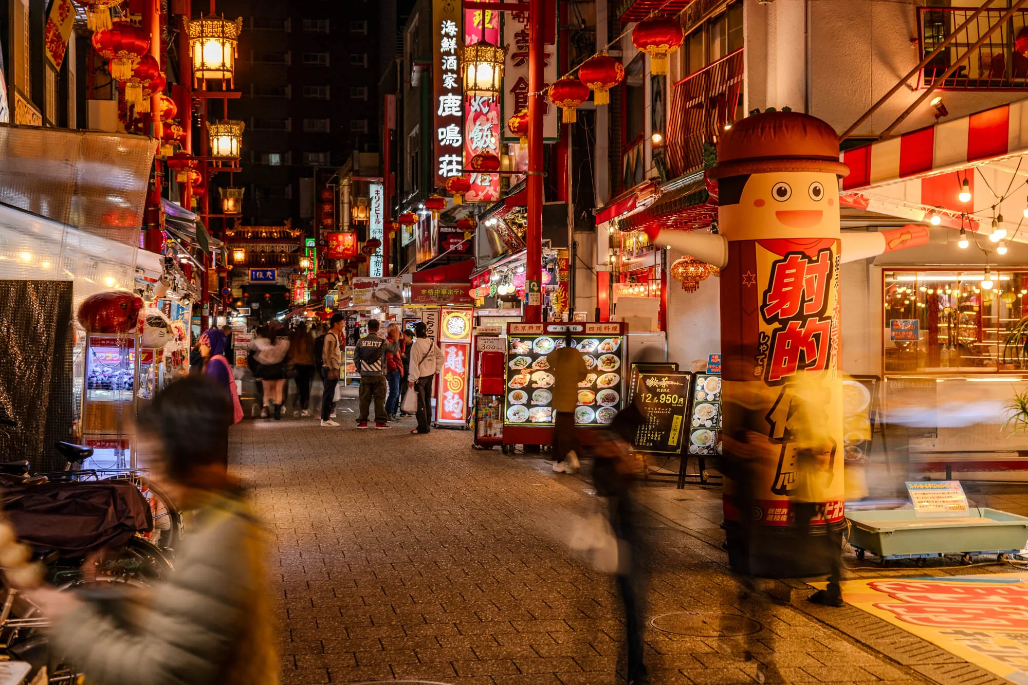 A vibrant night scene depicts a bustling street within Kobe Chinatown, illuminated by countless red and yellow lanterns and bright neon signs. Pedestrians move through the narrow, cobblestone-like thoroughfare, their forms blurred into dynamic streaks of color and light. A large, cheerful cartoon mascot figure, predominantly red and yellow with Japanese text, stands prominently on the right side of the street. Storefronts displaying food and other goods line both sides, receding into a distant glow where more people gather under festive lights.