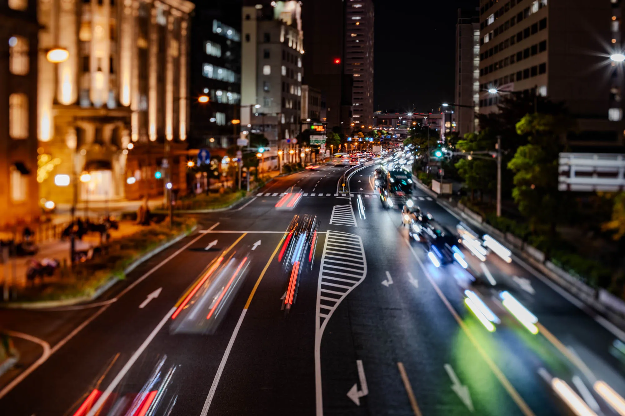 A dynamic, high-angle view captures a bustling city intersection at night, illuminated by the glow of streetlights and architectural lighting. Long exposure photography transforms moving vehicles into vibrant streaks of red taillights and cool white headlights, flowing across the multi-lane asphalt road. Tall, modern buildings line the right side, while a grand, ornate structure with warm-toned lighting stands on the left. White lane markings and crosswalks define the complex road network, directing the blurred motion of traffic. The scene's tilt-shift effect creates a selective focus, emphasizing the central traffic flow while blurring the periphery, lending a sense of miniature scale to the urban landscape.