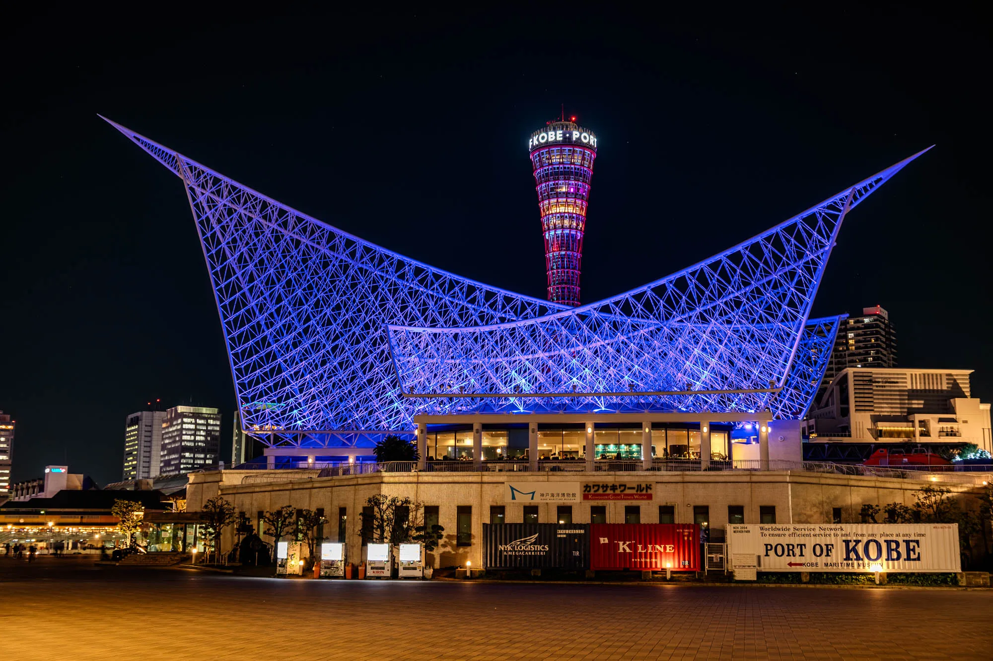 A vibrant night photograph captures the illuminated Kobe Port skyline. A striking white lattice structure, glowing intensely with electric blue light, dominates the foreground, its sweeping wings framing a slender, spiral-shaped tower behind it. The tower, emblazoned with "KOBE PORT" at its peak, radiates a gradient of warm red, pink, and purple light, contrasting with the cool blue of the lattice. Below the grand structures, a paved plaza, warmly lit, leads to a building with visible windows and, further right, two large shipping containers, one clearly marked "PORT OF KOBE." The long exposure technique renders the architectural lights with vivid intensity against the deep black night sky.