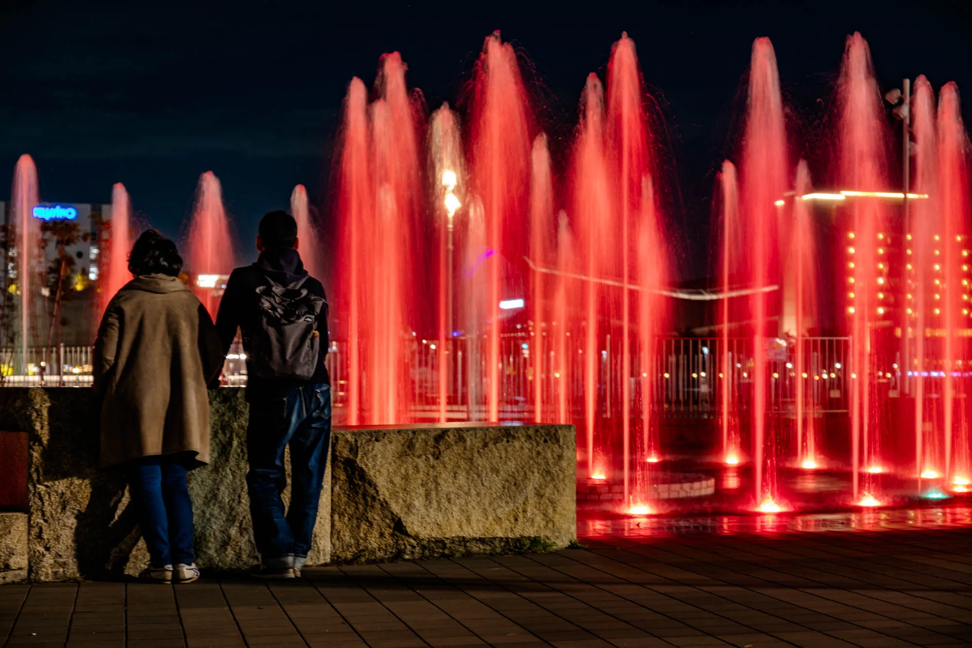A man and a woman, seen from behind, stand hand-in-hand before an array of intensely red-illuminated water fountains at night. The woman wears a light brown coat and blue jeans, while the man sports a dark hoodie and backpack, both gazing forward. A low, rough-hewn stone wall separates them from the vibrant aquatic display. The long exposure renders the cascading water into smooth, ethereal pillars of crimson light, creating a dramatic curtain against the deep blue-black sky. The paved ground beneath them subtly reflects the fiery glow.