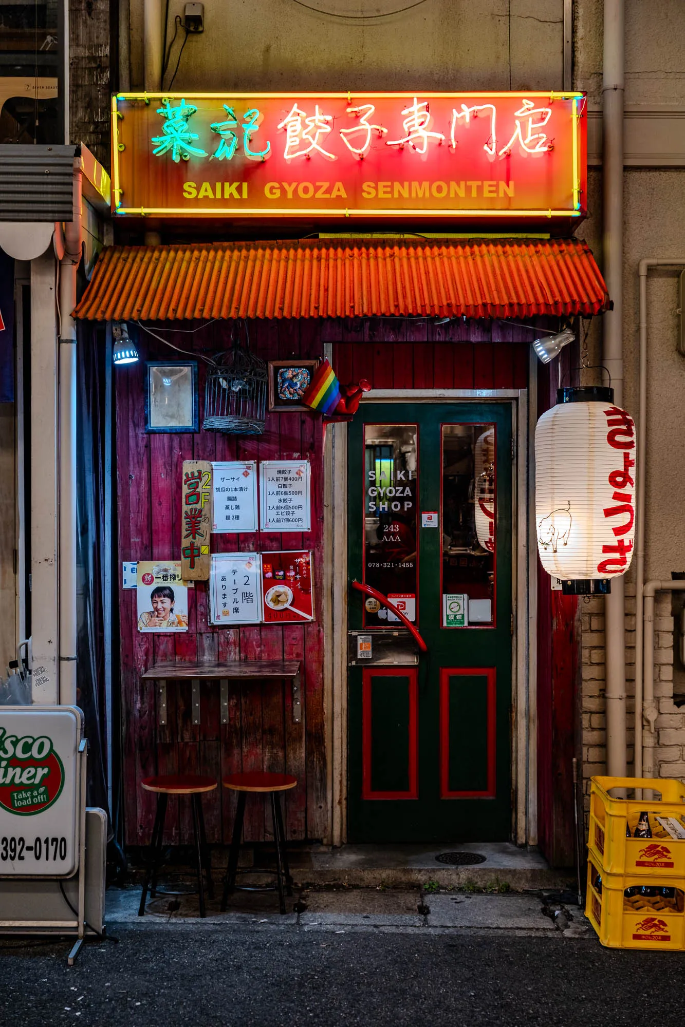 A vibrant, narrow storefront glows with neon lights and eclectic decorations at night. Above a textured orange awning, a large rectangular sign displays Japanese characters in bright green and red neon, alongside "SAIKI GYOZA SENMONTEN." The shop's weathered red wooden facade features a dark green door with red-trimmed panels, flanked by various posters, framed images, and a small, inviting outdoor counter with two red stools. To the right, a large white paper lantern, adorned with red script and a pig illustration, casts a warm glow beside stacked yellow crates of bottles, adding to the bustling urban atmosphere.