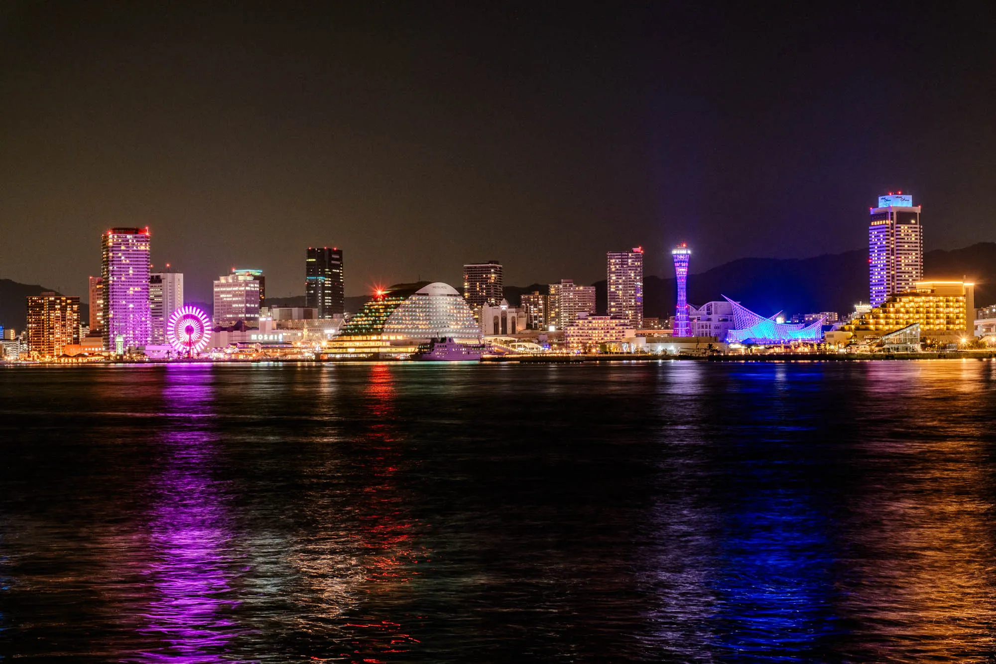 A long-exposure panoramic night view reveals a vibrant urban waterfront, its diverse architecture brilliantly illuminated and reflected across a body of water. On the left, a towering magenta-lit Ferris wheel punctuates a cluster of tall buildings glowing with purple and white lights. Towards the center, a striking pyramid-shaped building with a terraced, illuminated roof sits beside a slender, latticework tower bathed in blue, along with a distinctive geometric white structure radiating bright blue light. The foreground water, smoothed by the long exposure, vividly mirrors the city's purple, red, and blue electric glow against the dark sky and distant hills.