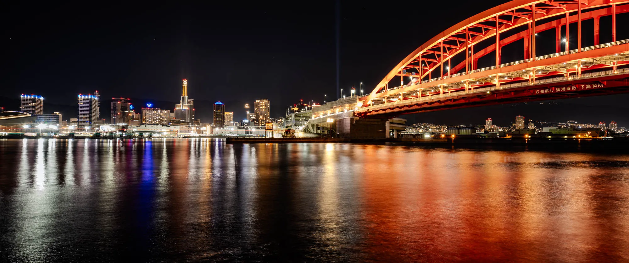 A panoramic night shot showcases the illuminated Kobe Ohashi Bridge and city skyline across a dark expanse of water. The vibrant red steel truss arch bridge dominates the right, its intricate structure brilliantly lit and reflecting a warm orange glow onto the rippled water below. To the left, a dense cluster of high-rise buildings glows with a spectrum of white, yellow, and blue lights, their reflections stretching across the water's surface due to a long exposure. The distant cityscape extends under a dark night sky, with faint stars visible above the mountain silhouettes.