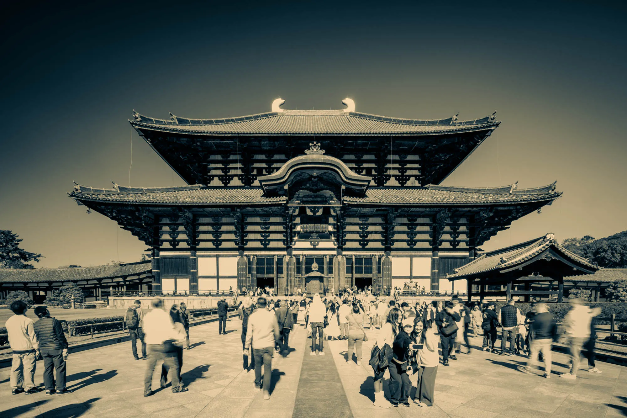 Todai-ji Temple, a monumental traditional Japanese Buddhist structure, stands prominently under a clear sky, its imposing dark tiered roofs and white facade dominating the midground. A bustling crowd of visitors fills the wide plaza in the foreground, many rendered with noticeable motion blur due to a slow shutter speed, capturing their movement across the stone ground. The entire scene is presented in a striking duotone, where deep blue-green tones articulate the sky and architectural shadows, while warm beige highlights the building's walls and the sunlit pavement, evoking a historical atmosphere.
