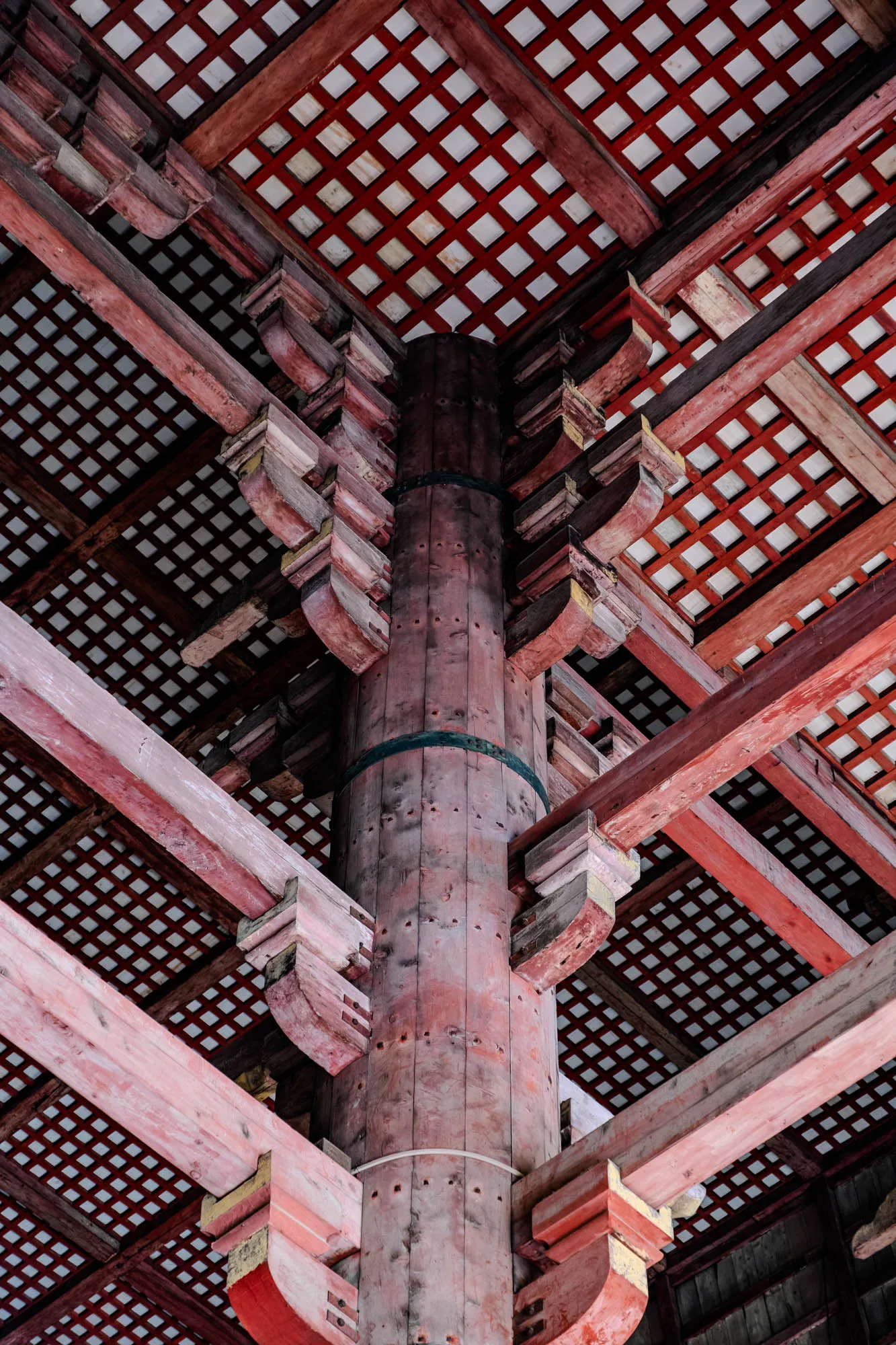 An intricate network of aged reddish-brown wooden beams and supports radiates from a large central pillar, forming the structural ceiling of a grand traditional building. The weathered central pillar, displaying distinct vertical wood grain and a subtle teal band, supports multiple layers of precisely carved and interlocking brackets. Above the complex joinery of the beams, the ceiling features a repetitive grid pattern of white squares set within a darker red wooden lattice. The low-angle perspective emphasizes the scale and meticulous craftsmanship of the architecture.