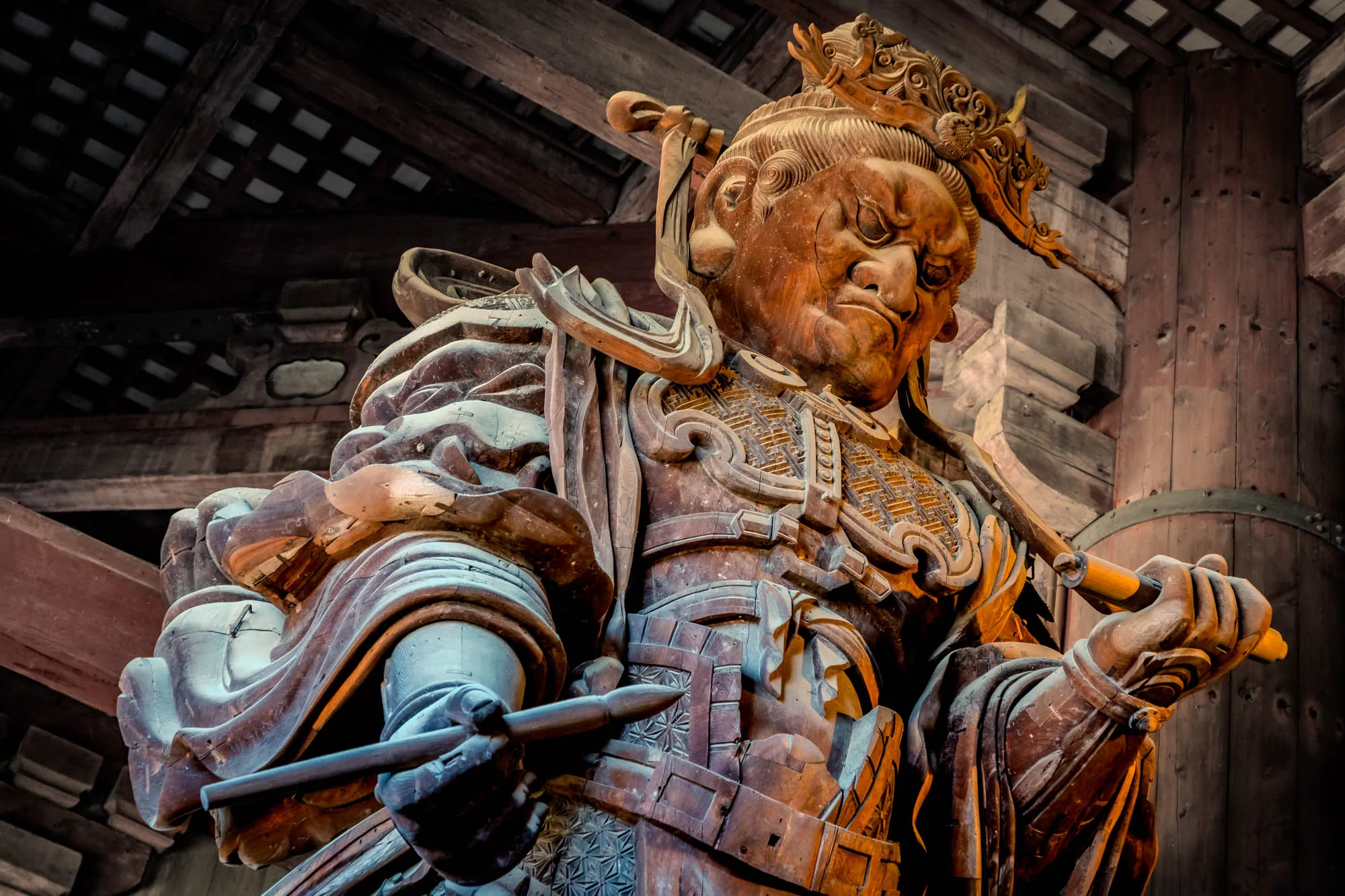 A towering, intricately carved wooden statue of a formidable guardian fills the low-angle frame, rendered in rich, aged wood. The figure wears an elaborate headdress and complex armor with geometric patterns, its stern face marked by deeply furrowed brows and a resolute gaze. Its powerful hands firmly grasp two staff-like implements, one held horizontally and the other angled upwards, emphasizing its commanding posture. The statue stands within a dimly lit interior, surrounded by dark, heavy wooden architectural elements including prominent beams and a distinct lattice ceiling.