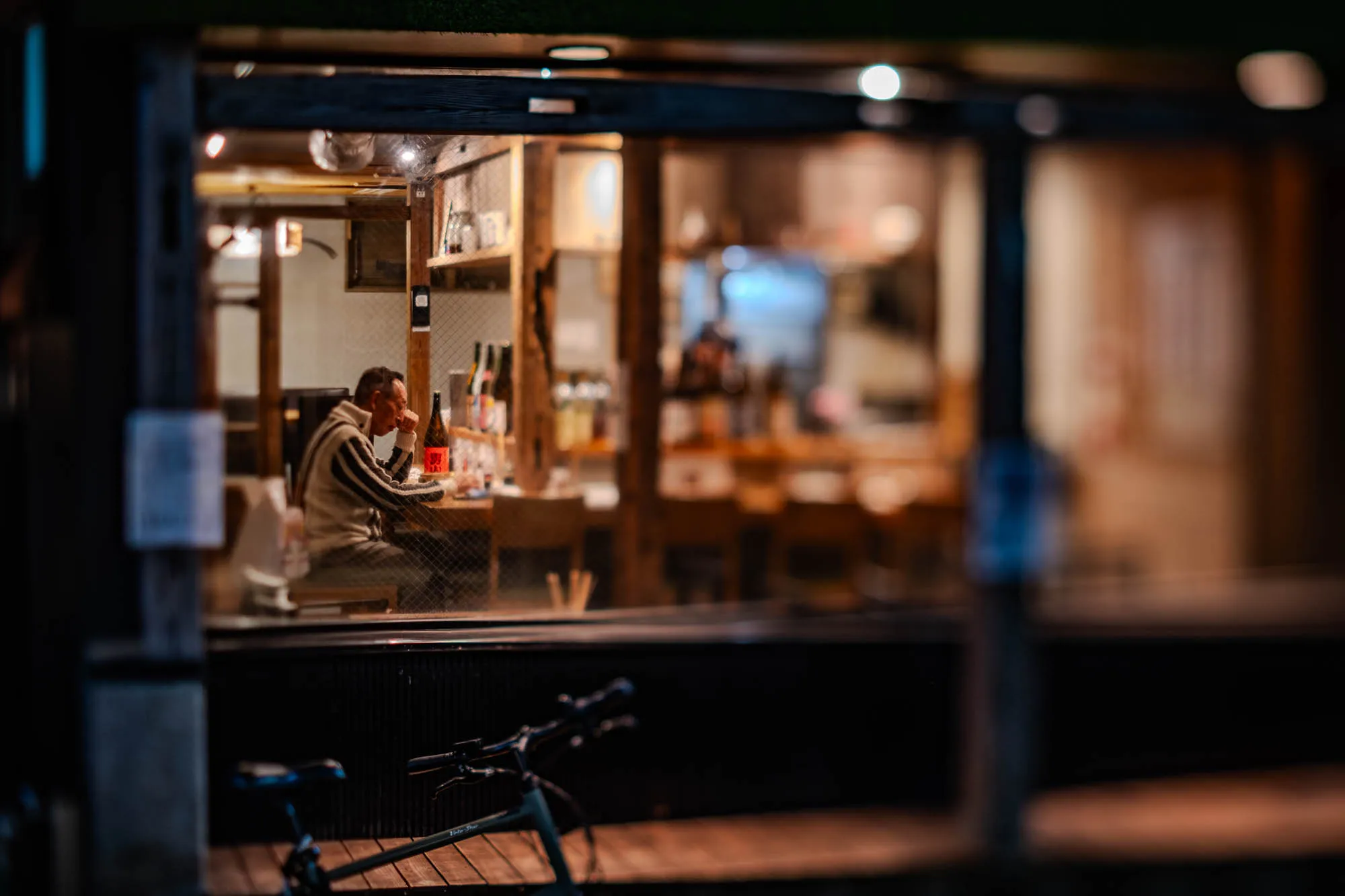 A man, seen through a large glass window, sits contemplatively at a wooden counter inside a warmly lit establishment at dusk. He wears a light-colored, striped sweater, with his right hand resting against his chin, his gaze directed slightly downwards. Bottles and various indistinct items line shelves behind him, rendered soft and blurry due to the shallow depth of field, which sharply isolates the man in focus. In the dark foreground, the handlebars and saddle of a bicycle rest out of focus, framed by the window's dark sill and frame.