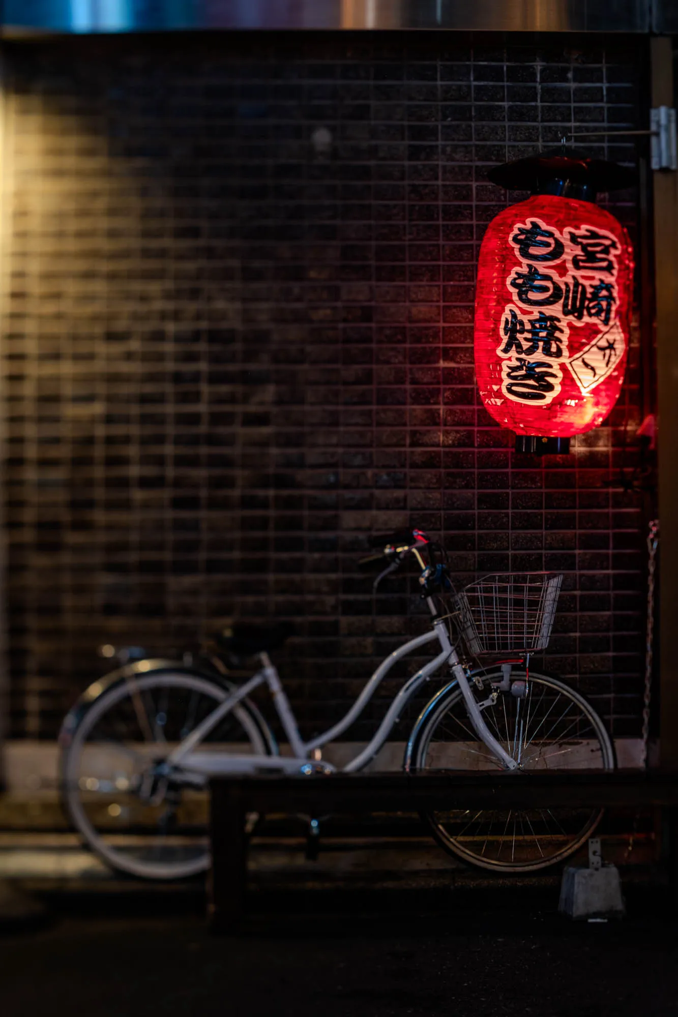 A vibrant red Japanese chōchin lantern, adorned with black calligraphy, hangs prominently on the right side of a dark, tiled building facade. Below it, a white step-through bicycle with a wire front basket is parked, its front wheel partially obscured by a dark, blurred wooden railing in the immediate foreground. The lantern's intense glow casts a warm red hue onto the textured wall, while a softer, yellowish light emanates from the far left, creating a subtle gradient. A very shallow depth of field keeps the lantern and the bicycle's front section sharply in focus, while the rest of the scene recedes into a soft blur.