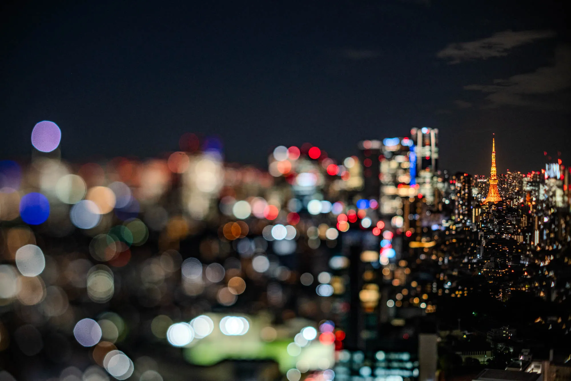 A vibrant night cityscape presents the illuminated orange Tokyo Tower in sharp focus, rising above a densely packed urban environment on the right. The foreground and left side are rendered in a dramatic blur of large, colorful bokeh circles, creating a dreamlike visual effect. This striking selective focus, achieved through a tilt-shift perspective, directs attention to the crisply detailed distant buildings and the iconic tower. Its warm glow stands in stark contrast to the soft, scattered lights of the immediate surroundings, all beneath a dark sky with faint clouds.