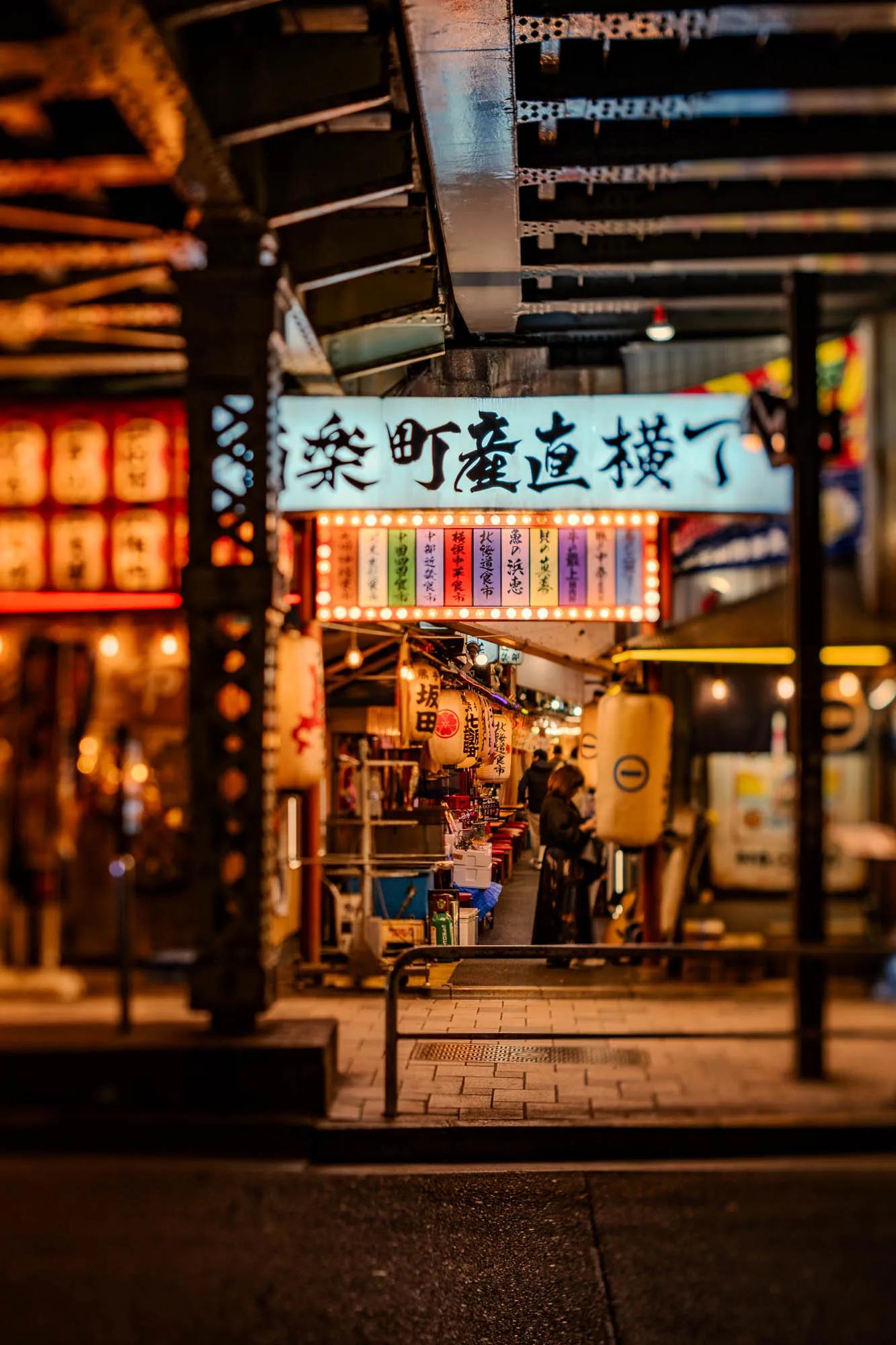 A brightly illuminated horizontal sign, adorned with bold Japanese characters, serves as a prominent gateway beneath the dark, riveted metal structure of an elevated track. The extremely shallow depth of field renders the dark foreground street and distant background as soft blurs, enhancing the vibrant focus on the bustling alleyway directly beyond the sign. Within this narrow passage, numerous hanging lanterns cast a warm glow over a pathway where patrons move amidst small, colorful stalls and storefronts. The scene captures the lively, nocturnal ambiance of an urban Japanese arcade, framed by the gritty industrial overhead.