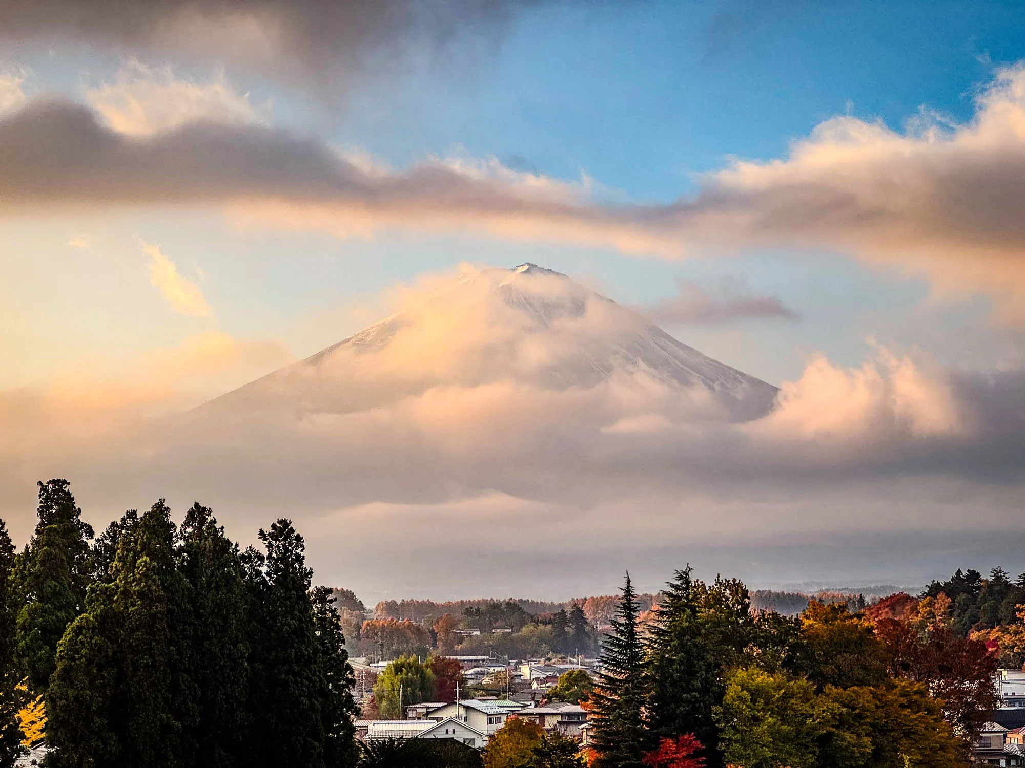 Mount Fuji's snow-capped peak emerges majestically from a dynamic shroud of clouds, illuminated by golden light against a clear blue sky. The lower slopes of the iconic volcano are softened by these atmospheric veils, which glow with warm hues from the rising sun. In the foreground, a vibrant landscape of dense evergreen trees and deciduous foliage in shades of red, orange, and yellow autumn colors densely frames a small town with various buildings.