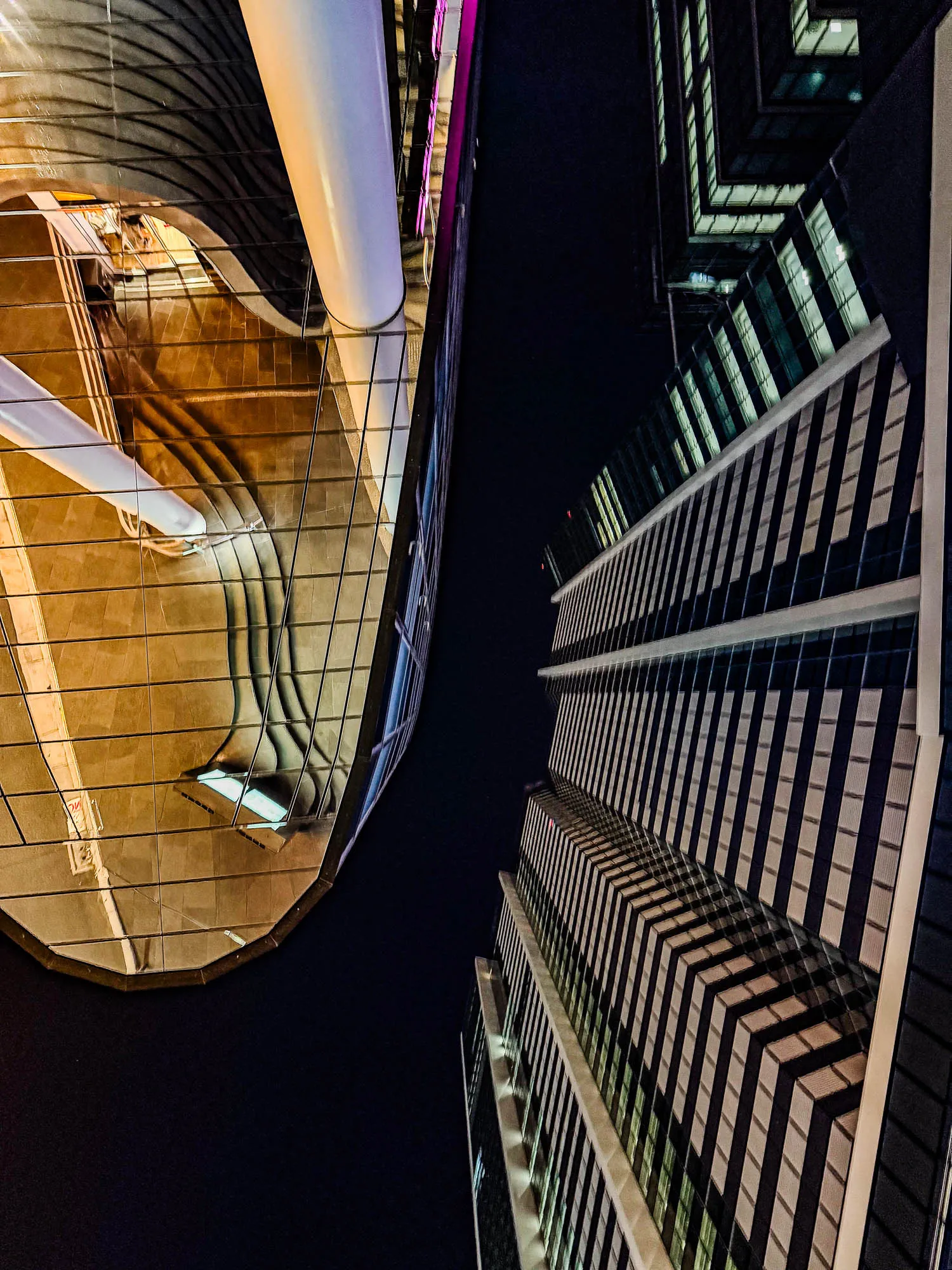 A dynamic low-angle view showcases two contrasting modern skyscrapers against a dark night sky. On the left, a building with a smoothly curved, gold-toned reflective facade dramatically mirrors the ground below it, interrupted by a prominent white cylindrical column. To the right, a taller, angular structure presents a rhythmic pattern of illuminated rectangular windows and dark panels, casting a cool, greenish glow. The sharp lines and deep shadows emphasize the architectural forms in this nocturnal urban scene.