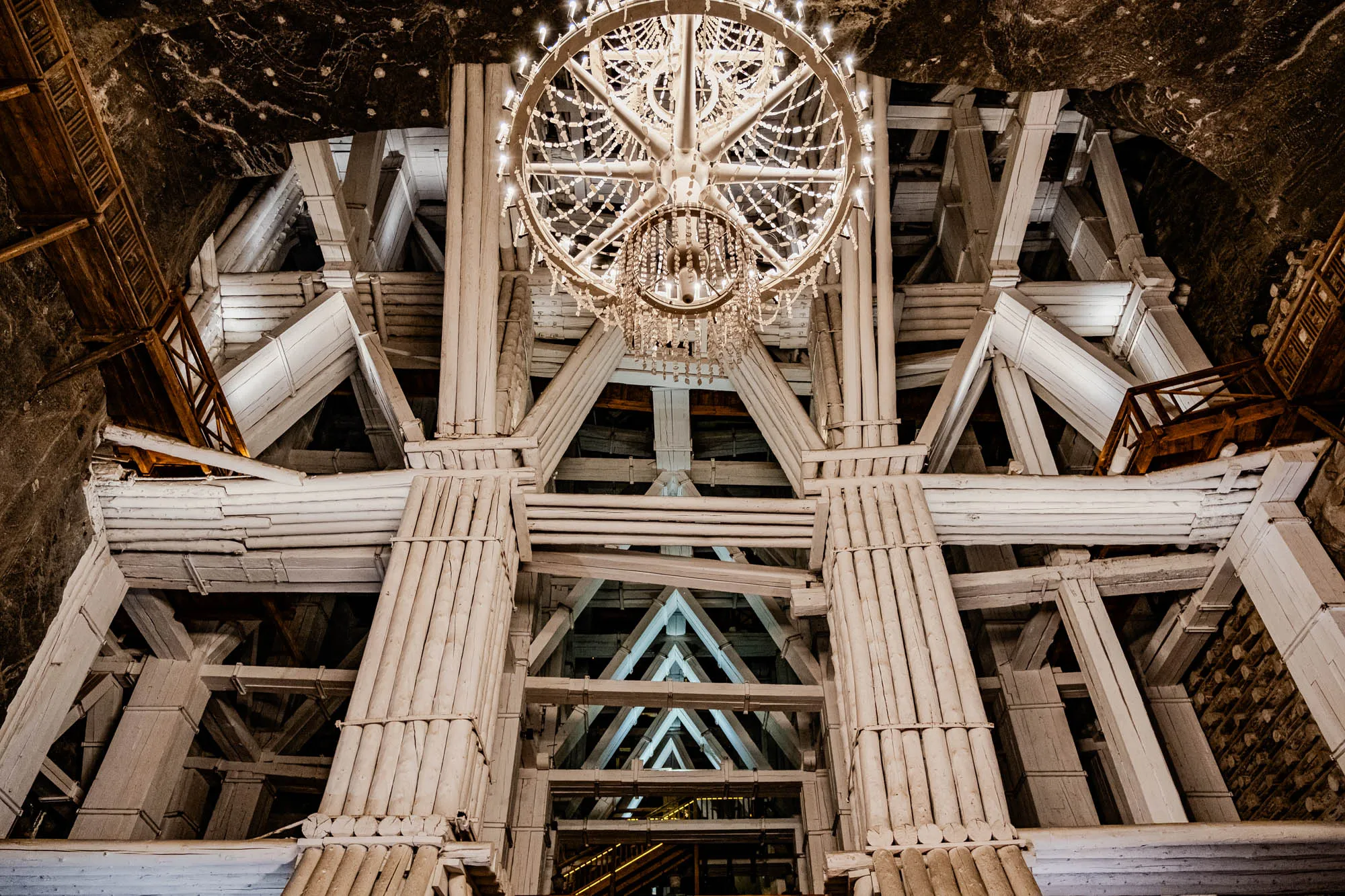 This image is of a large, ornate chandelier suspended from a high ceiling. The chandelier is made of metal and glass, with a circular frame and numerous crystal pendants hanging from it.  The chandelier is illuminated by several light bulbs, and its reflection can be seen in the surrounding surfaces. The ceiling itself is constructed from a complex network of wooden beams, arranged in a pattern of triangles and squares. The beams are white, and they cast long shadows on the floor. The image is taken from a low angle, looking up at the chandelier and ceiling. It's reminiscent of a cathedral, suggesting an underground chamber or a historic salt mine. The dark stone ceiling above the beams suggests a space that was once a natural cave or tunnel.