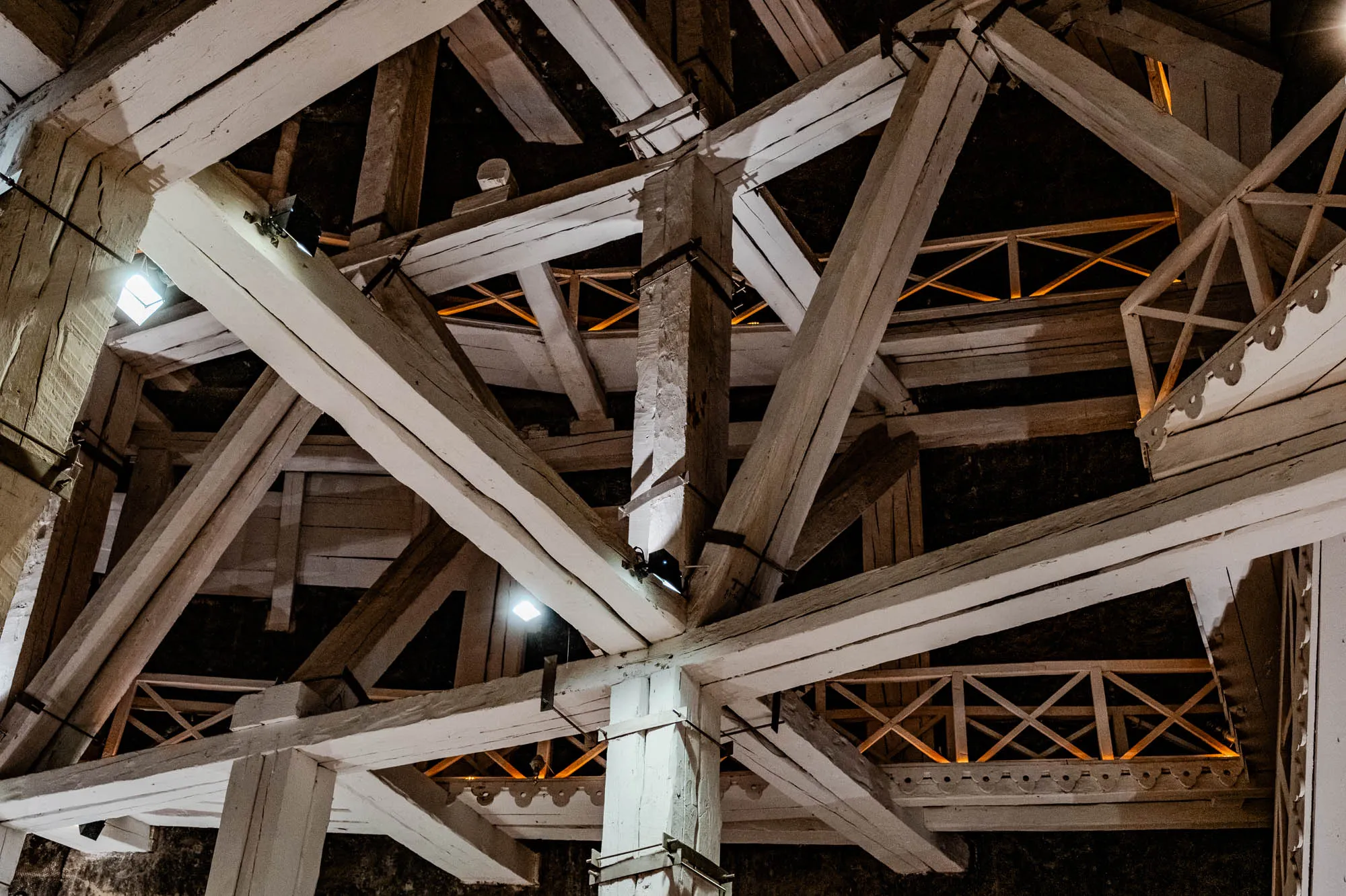 The image shows a close up of the interior of a building with a very complex wooden structure. The structure appears to be a roof truss, with many beams and supports criss-crossing to form a complex network. The beams are white and painted, with some showing the natural wood grain. The supports are large and solid, and they create a sense of strength and stability. The image is taken from a low angle, looking up at the structure. There are three light sources visible, casting warm light on the wood.