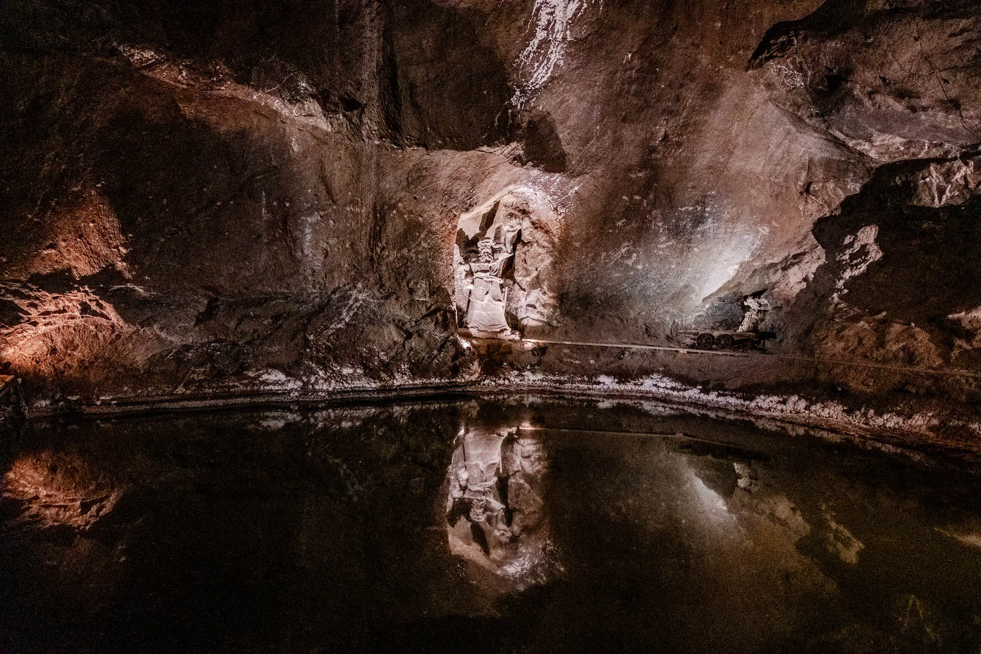 The image shows a dark, rocky cave with a pool of water in the center. The cave ceiling and walls are rough and uneven, with the texture of rock and some white salt deposits. The pool of water reflects the cave's features, including a small opening to another part of the cave, and the light from an unknown source. It appears as if the cave is lit by artificial light, casting a dim glow on the rock and water. The atmosphere is one of mystery and stillness, with the cave seemingly untouched by human activity.