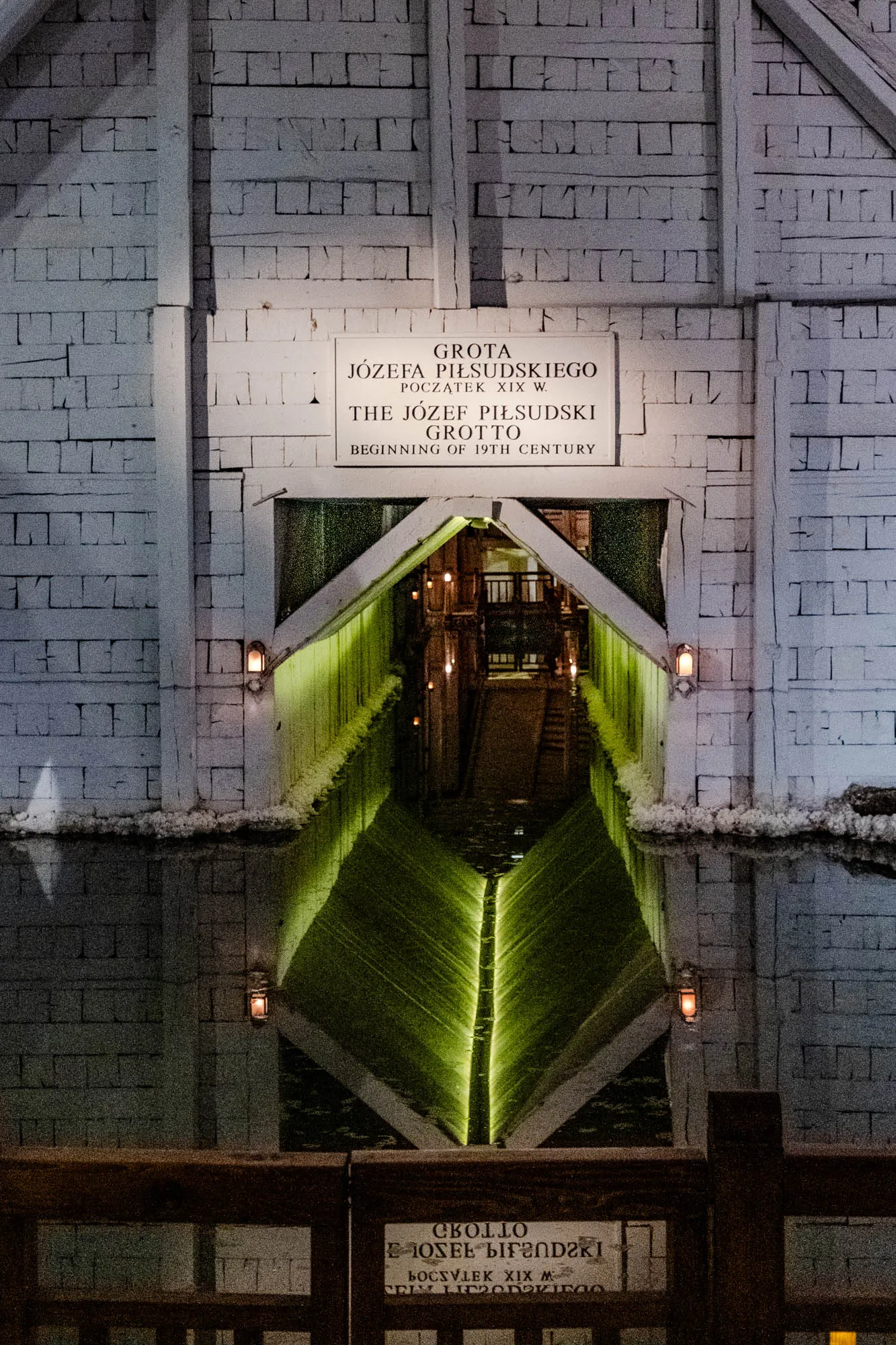 The image shows a narrow, dark tunnel made of white brick. It appears to be a passageway through a stone wall.  A sign above the entrance is inscribed with the words "GROTA JÓZEFA PIŁSUDSKIEGO POCZĄTEK XIX W. THE JÓZEF PIŁSUDSKI GROTTO BEGINNING OF 19TH CENTURY" in English and Polish. The tunnel itself is mostly dark except for a band of green light in the middle, which is reflected in a pool of water at the bottom.  There are small, dimly lit lanterns lining the sides of the tunnel.  At the very bottom of the image, a wooden fence and another sign with the text "CROLLO BOCSVLEK XIX M" in Polish are visible.  This image depicts a tunnel entrance with a reflective pool of water and green light. 
