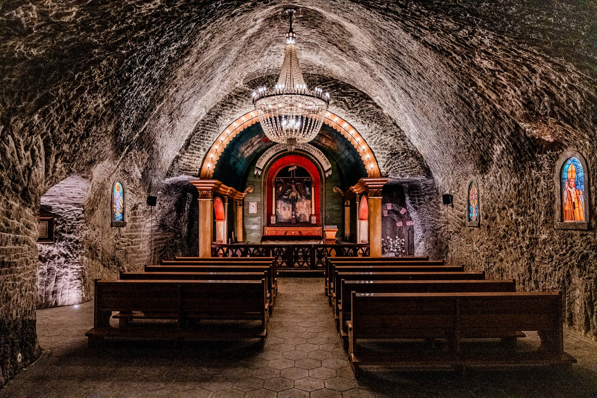 The image shows a chapel built into a salt mine. The chapel is dimly lit and has a vaulted ceiling made of salt rock. There is a large chandelier hanging from the center of the ceiling. The chapel has wooden pews arranged in rows facing a small altar at the far end. The altar has a stained-glass window depicting a religious scene. There are other stained-glass windows on the side walls of the chapel. The walls are made of rough salt rock and are uneven. The chapel is very dark and the light from the chandelier and windows creates a dramatic effect.