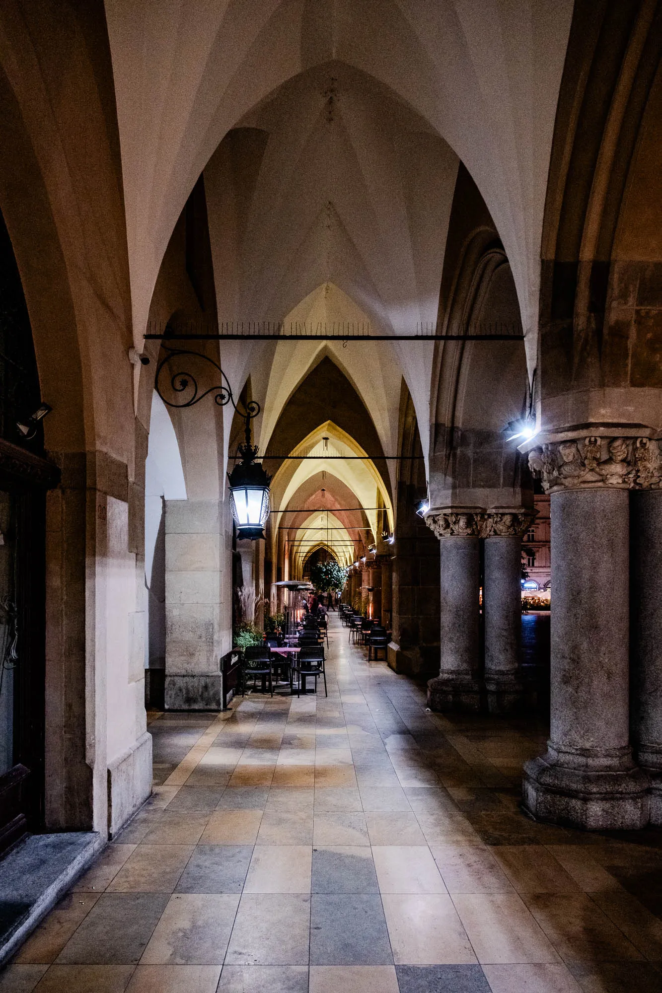 The image shows a long, narrow, stone-arched passageway. The passageway is lit by several overhead lights and features a series of pillars on each side. The pillars are made of a rough-hewn stone and some have decorative carvings. There are black metal railings on the sides of the passageway. The passageway leads to a slightly lighter area in the distance where there are some small tables and chairs. The floor of the passageway is made of large, square, stone tiles. The passageway's walls and the underside of the arch are made of a smooth, light gray stone. The arch is made of a thick, rough-hewn stone that is darker in color. The hallway is dark and somewhat mysterious.