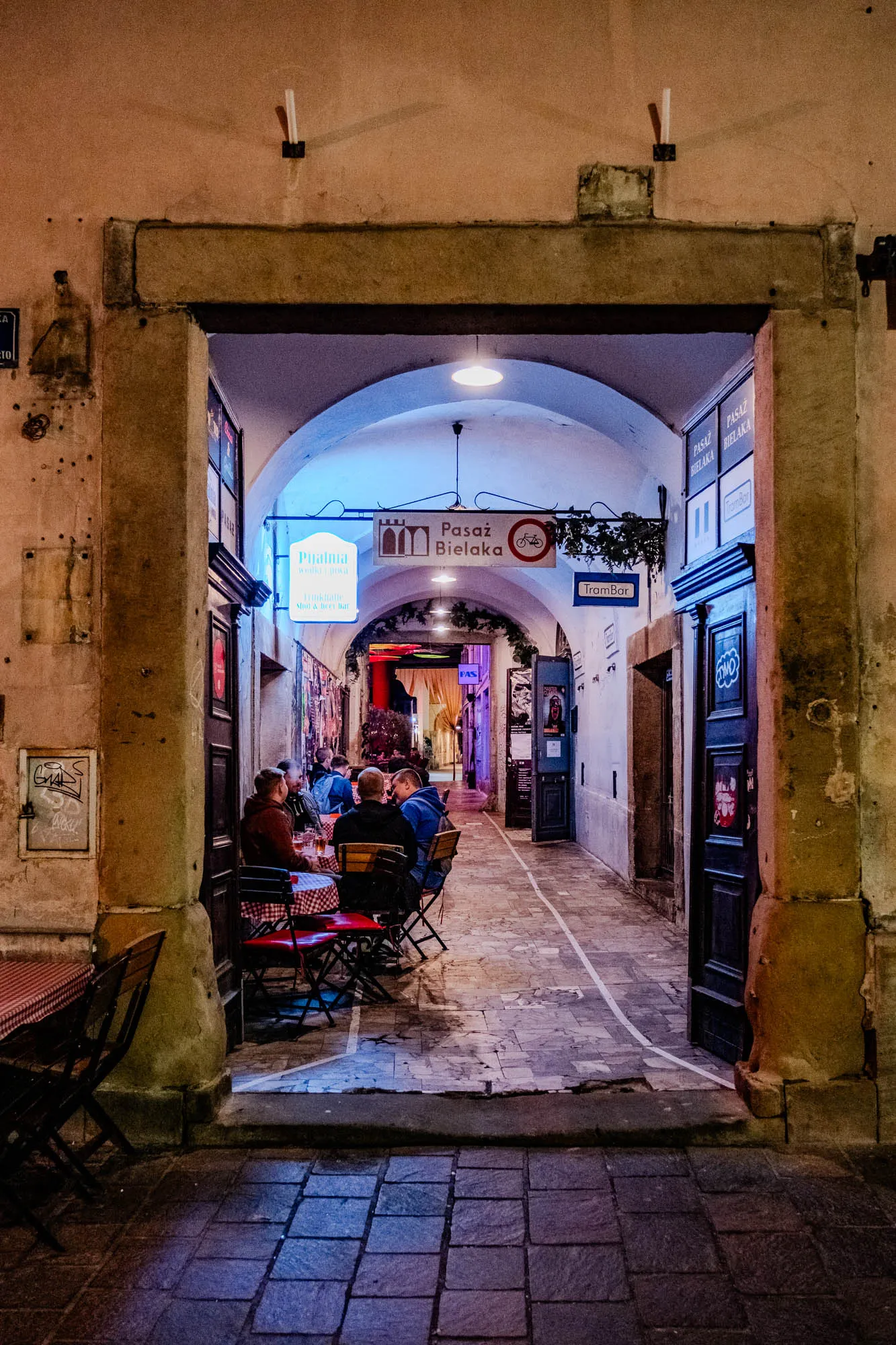 The image shows a narrow, arched passageway with a square opening lined with buildings on either side. It is lit by overhead lights and appears to be a pedestrian-only zone. 

On the left, several people are sitting at a table with red chairs and a checkered red and white tablecloth. Behind them is a door with a sign that reads "Pualnia wodki prwa Shot & beet Dar". To the right, the passageway opens up into an area with a sign that reads "Pasaż Bielaka". There is a sign that reads "TramBar" and a white line that appears to be a pedestrian walkway. To the right of the walkway is a door with a sign that reads "PASAZ BIELAKA". The image is taken at night and the buildings are all lit up, creating a warm and inviting atmosphere.