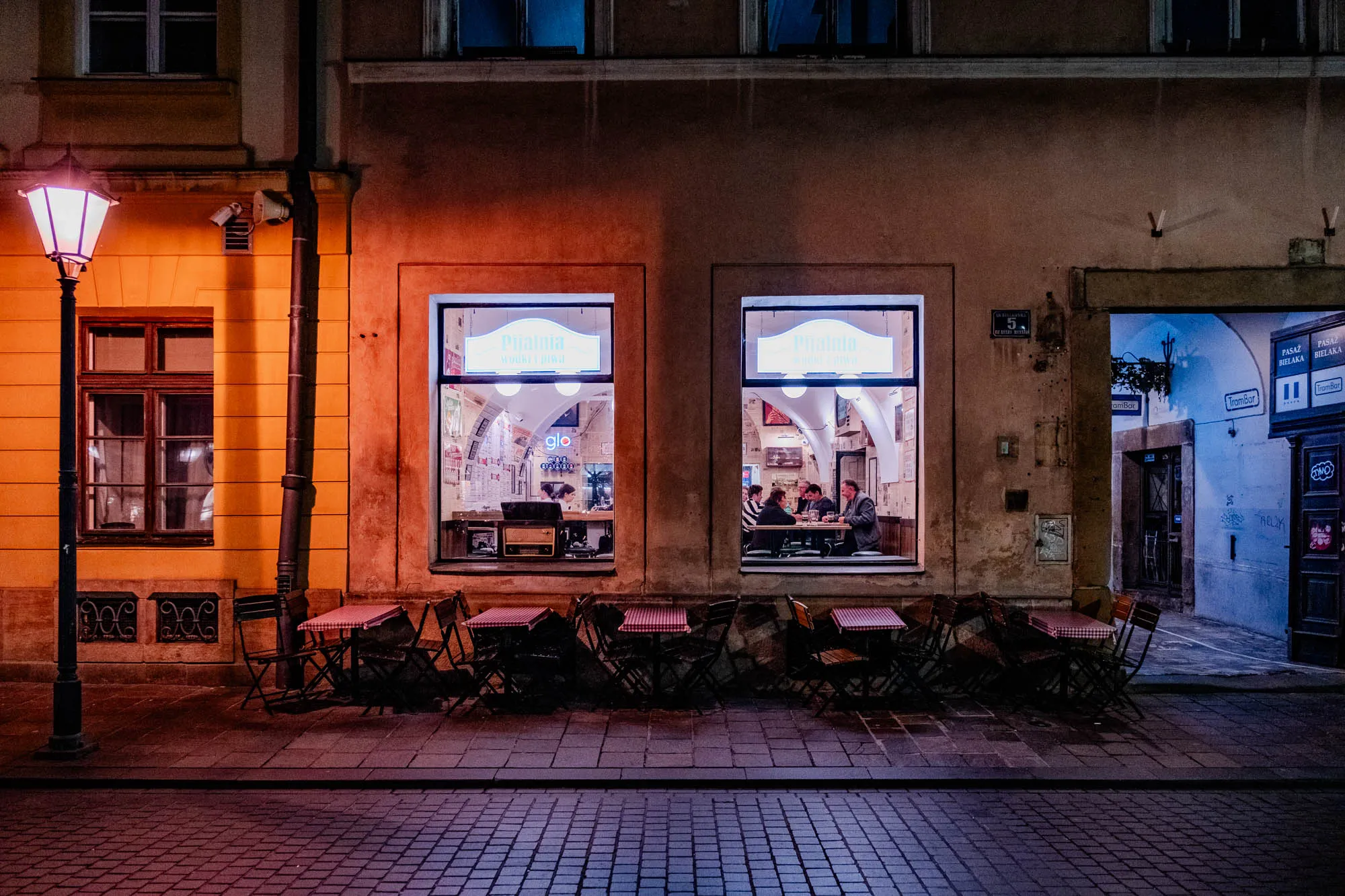 The image depicts a street scene at night. On the left, a bright yellow building with a lit street lamp stands. The lamppost is tall and black with a round, white globe light. In front of the building are a few small, black metal chairs and tables. On the right is another building, lit up by a warm yellow glow. There are two large windows with glass panes that display the interior of the shop. Inside the windows, there are people sitting at tables and a neon sign that reads "Pijalnia Wodkryjiwa."  There are also several black, metal chairs and tables outside this building. The street between the buildings is cobblestone and stretches off into the distance.  In the background, on the right side of the image, a small alleyway can be seen with a blue wall. This wall has some writing on it and a small sign that reads "TramBar".  There is a blue door at the end of the alleyway.  The image is lit by a warm glow from the streetlight and the windows. It is a peaceful, quiet scene with no one walking or driving by.