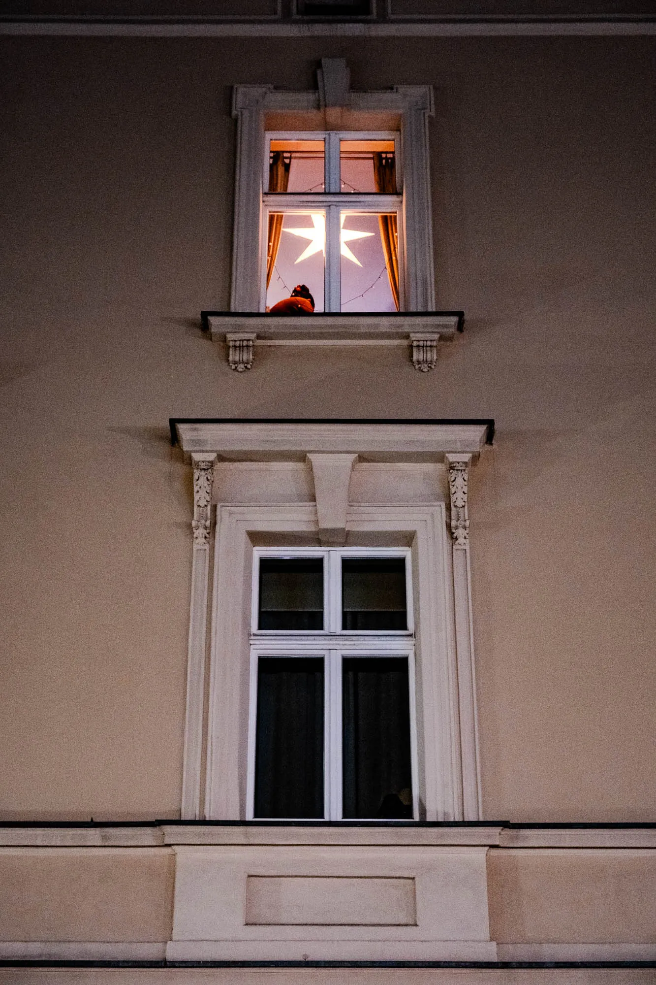 The image shows a beige building with two windows. The top window is lit and has a white star hanging in the window. You can see a figure in the window.  The bottom window is dark and appears to be closed.  The windows have decorative trim around them.