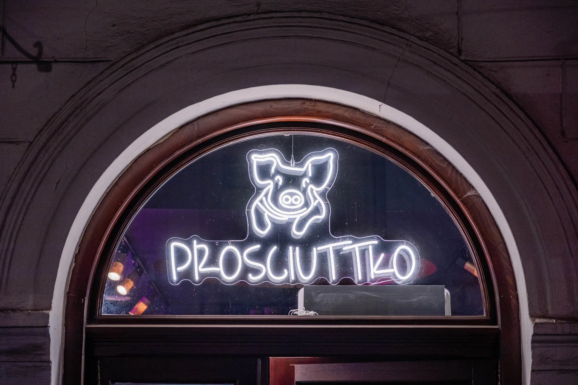 The image is of a storefront with an arched window. In the center of the window is a white neon sign that says "Prosciutto" in Polish in all caps. Above the text is a white neon pig's face. The pig has a slight smile and two ovals for eyes. The sign is in a dark window, with a blurry purple and blue background. The window is framed by a dark wood trim. The storefront is made of textured brick and has a white painted edge. The brick has a few cracks in it.  