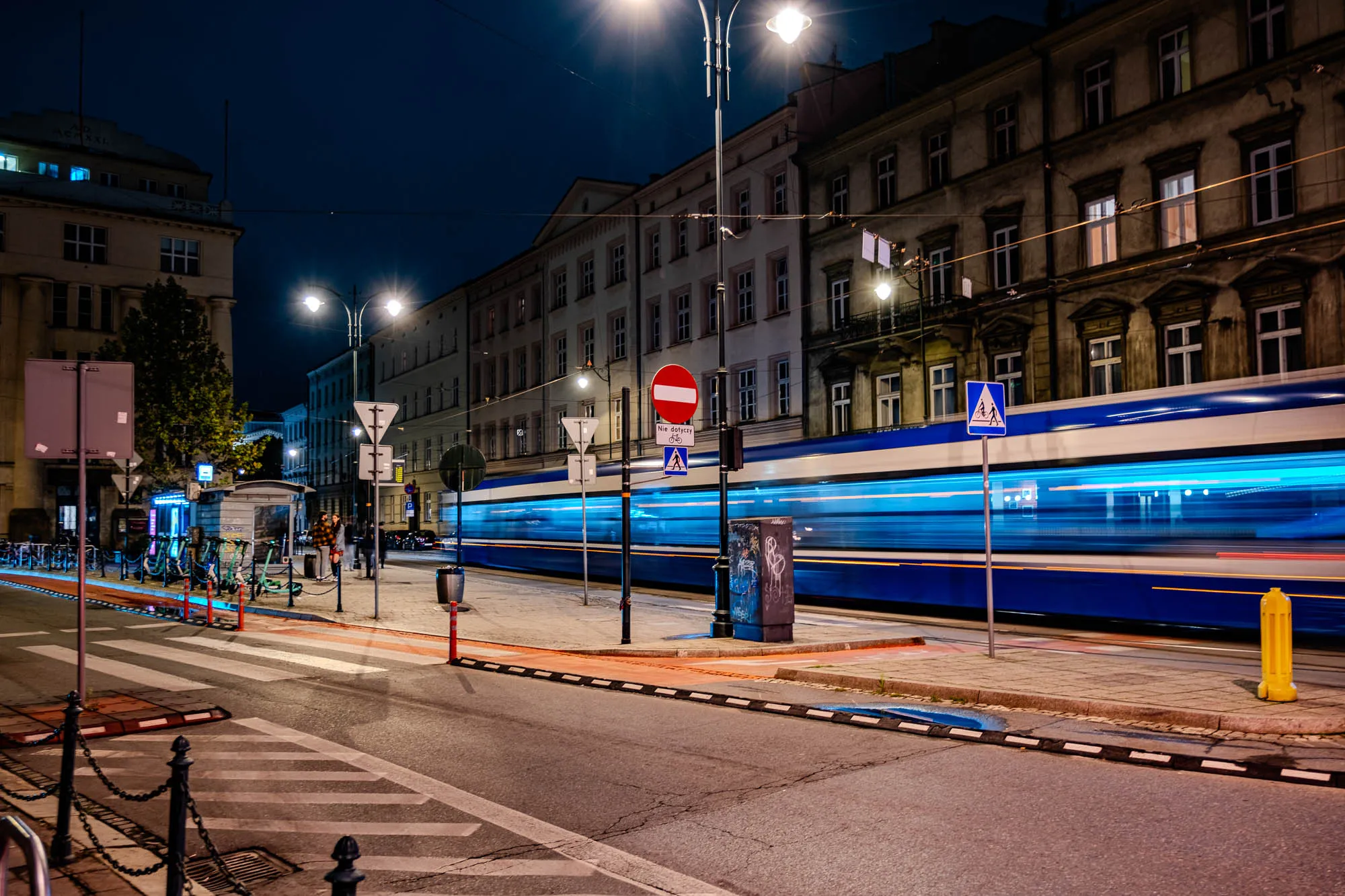 The image is a night shot of a street in a city. There is a streetlight in the center of the image that illuminates the street, a crosswalk, and a sidewalk. On the sidewalk are a row of bicycles, and two people standing by them. To the right of the sidewalk, a tram is moving quickly, its motion blurred in the image. Behind the tram, several tall buildings rise up, with streetlights shining down. The street has a black iron fence along the left side, a curb, and a cobbled sidewalk next to the tram. The buildings are all lit up and there are a few street signs in the image. The tram is a light blue and white color.  A streetlight shines onto the crosswalk, which has a white pedestrian crossing sign.  The streetlight is connected to a pole, that also holds a stop sign with a bicycle symbol and the text "Nie dotyczy" on it.  Next to the stop sign, a pedestrian crossing sign with a symbol of a person walking is visible.  A yellow fire hydrant can be seen to the right of the image.