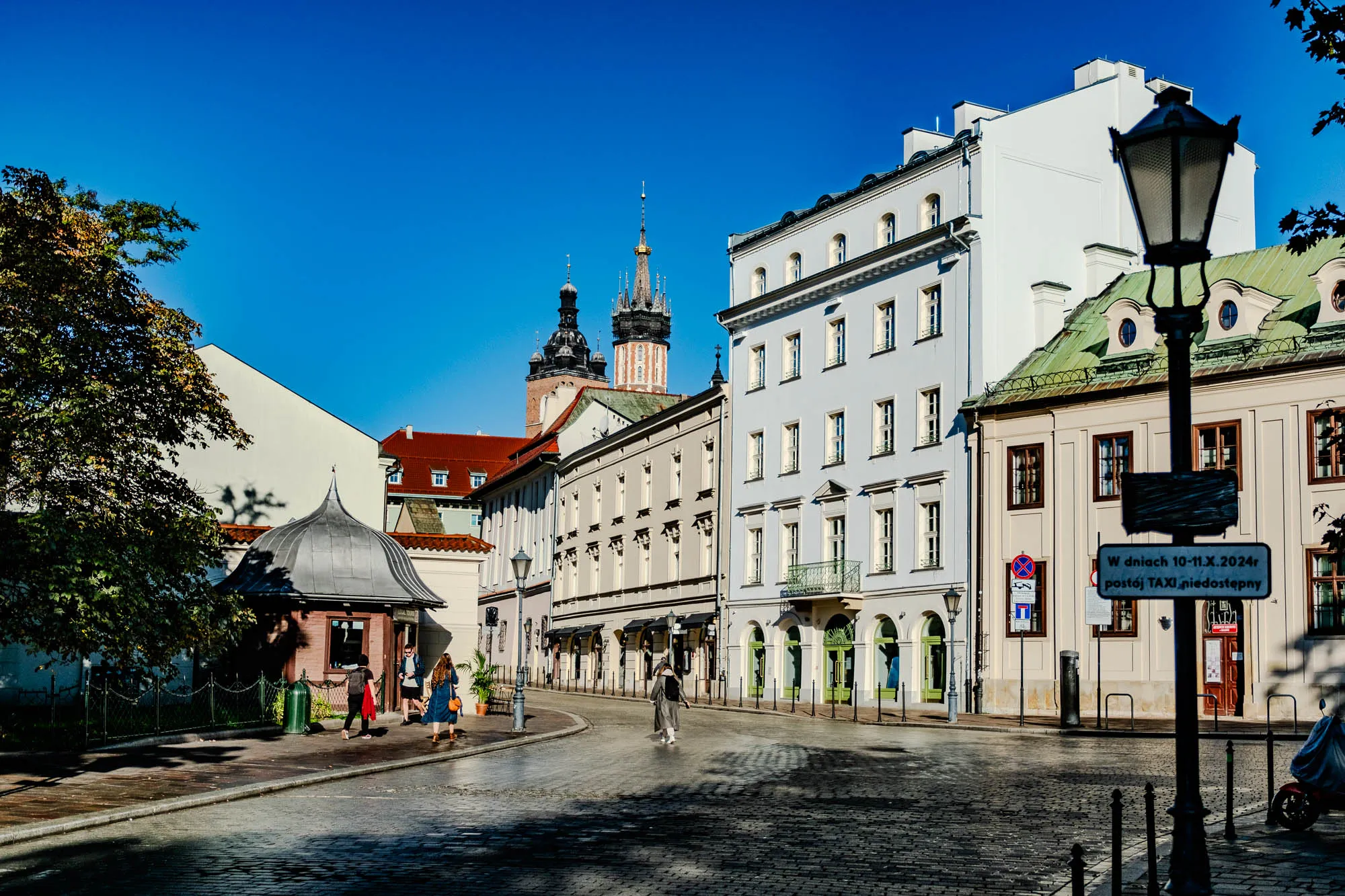 The image shows a cobblestone street in a European city. There are several buildings lining the street, all with white facades and dark-colored roofs. In the distance, two tall church towers can be seen, one with a golden spire. On the left side of the street, there is a large green tree and a small brown kiosk with a domed roof. Two women are walking in front of the kiosk. There is also a street lamp with a sign in Polish that says "In days 10-11.X.2024r. Taxi stand unavailable." The street is mostly empty and there is a blue sky in the background.