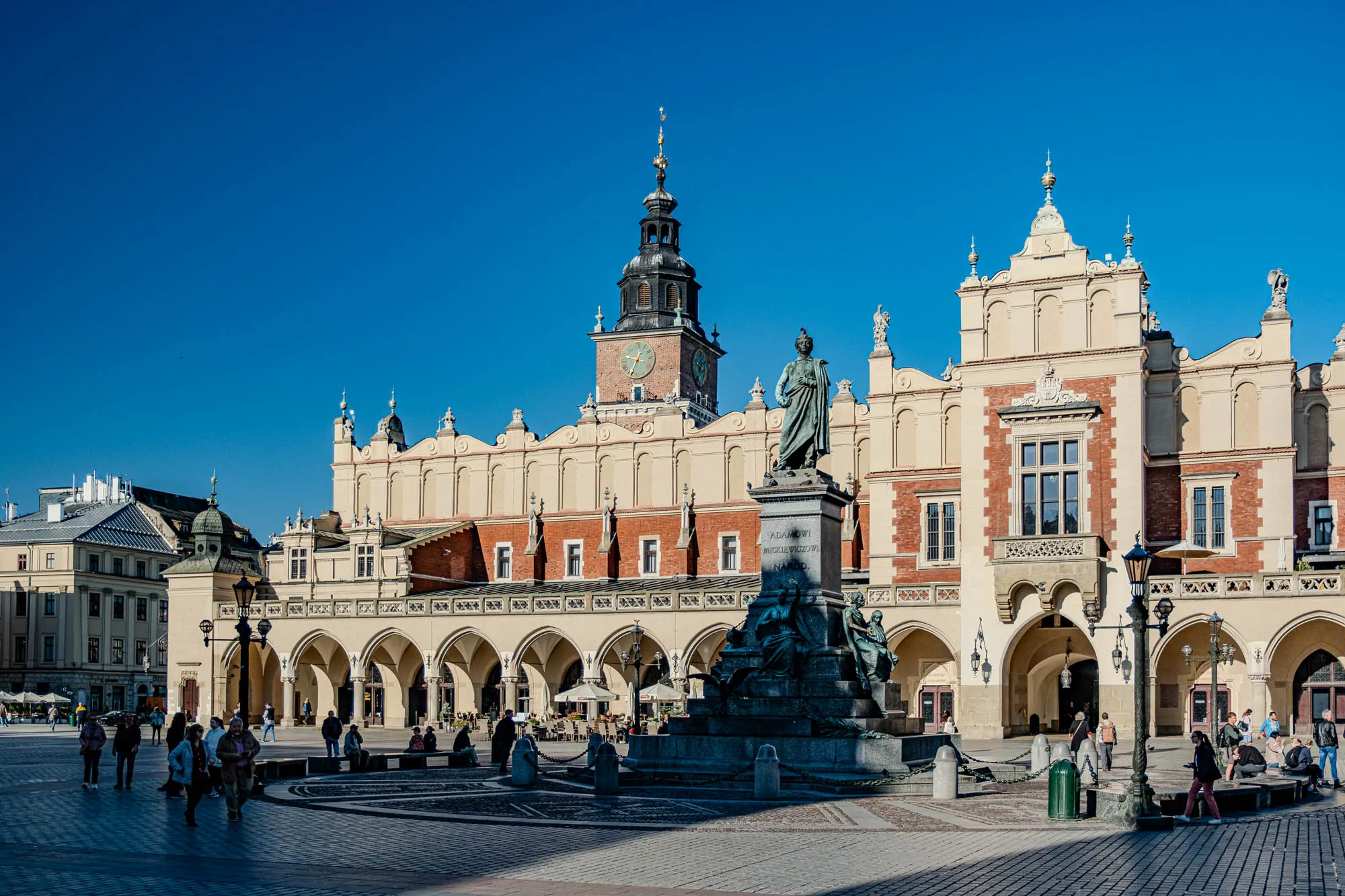 A large square in front of a historic building complex. The square is made of large cobblestones and is mostly empty. There are a few people walking around, and a few people sitting down. In the center of the square is a large bronze statue. The statue is of a man standing on a plinth and holding a scroll. The statue is surrounded by a low wall and some greenery. In the background there are a few street lamps and a building complex. The building has a high bell tower with a clock face on one of its sides. The building is light beige with red brick accents. The complex has multiple arches, and an ornate facade. The sky is bright blue and clear.