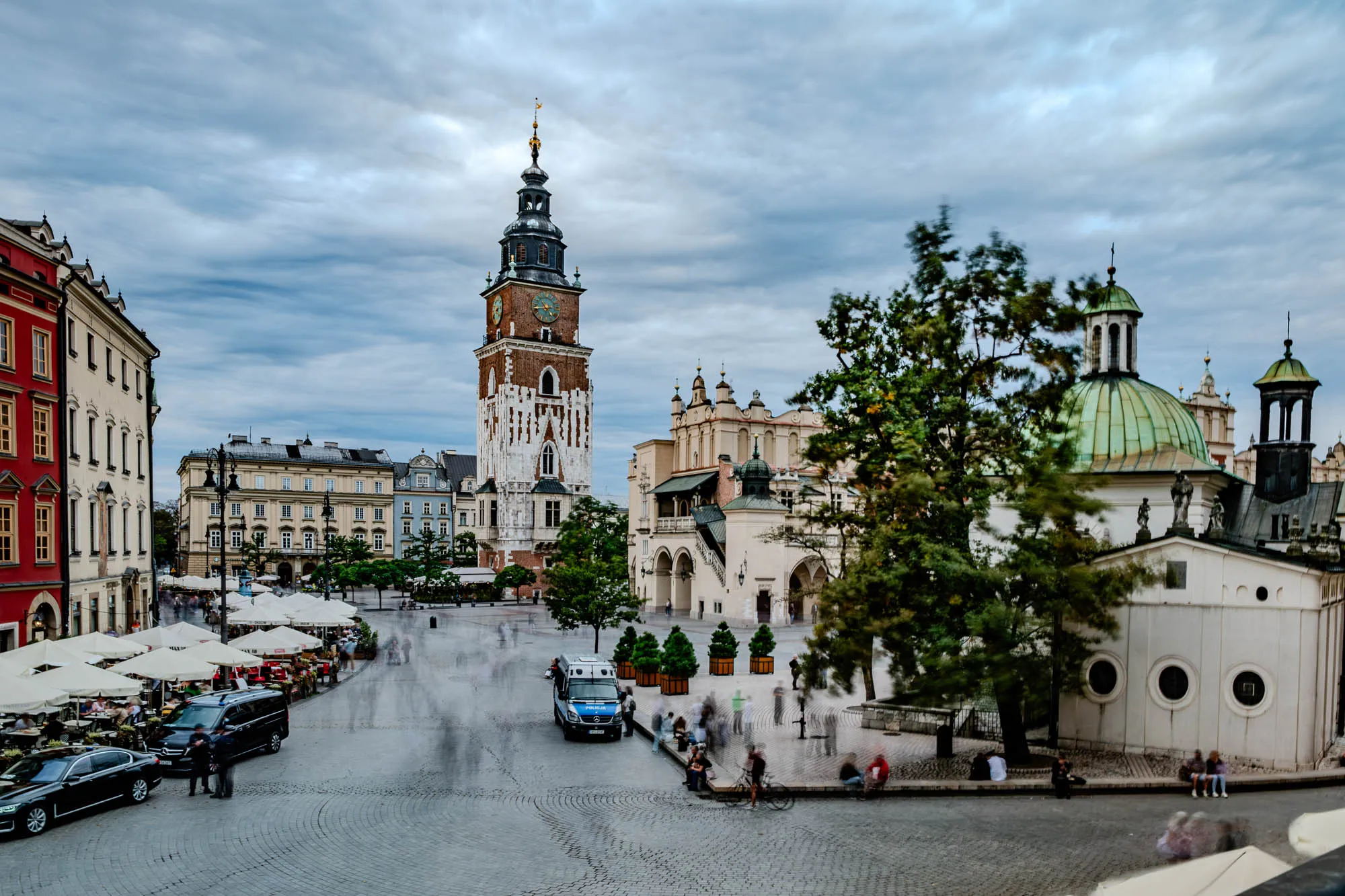 The image shows a large, cobblestone square in a European city. In the center of the square is a tall clock tower, with a pointed roof and a white and red facade. The tower is flanked on either side by a row of buildings, most of which are white and red with many windows. The buildings have a more modern style than the tower. The square is filled with people, many of whom are blurred because they are moving. There are also several white umbrellas lining the edge of the square, suggesting that there are cafes or restaurants in the area. Behind the tower and the buildings, there is a large tree with green leaves. The tree is in the foreground of the image and partially obscures a building with a green roof. The square is under a cloudy sky. The sky is a light blue color with white clouds. The clouds are thick and moving, and they look like a stormy sky.