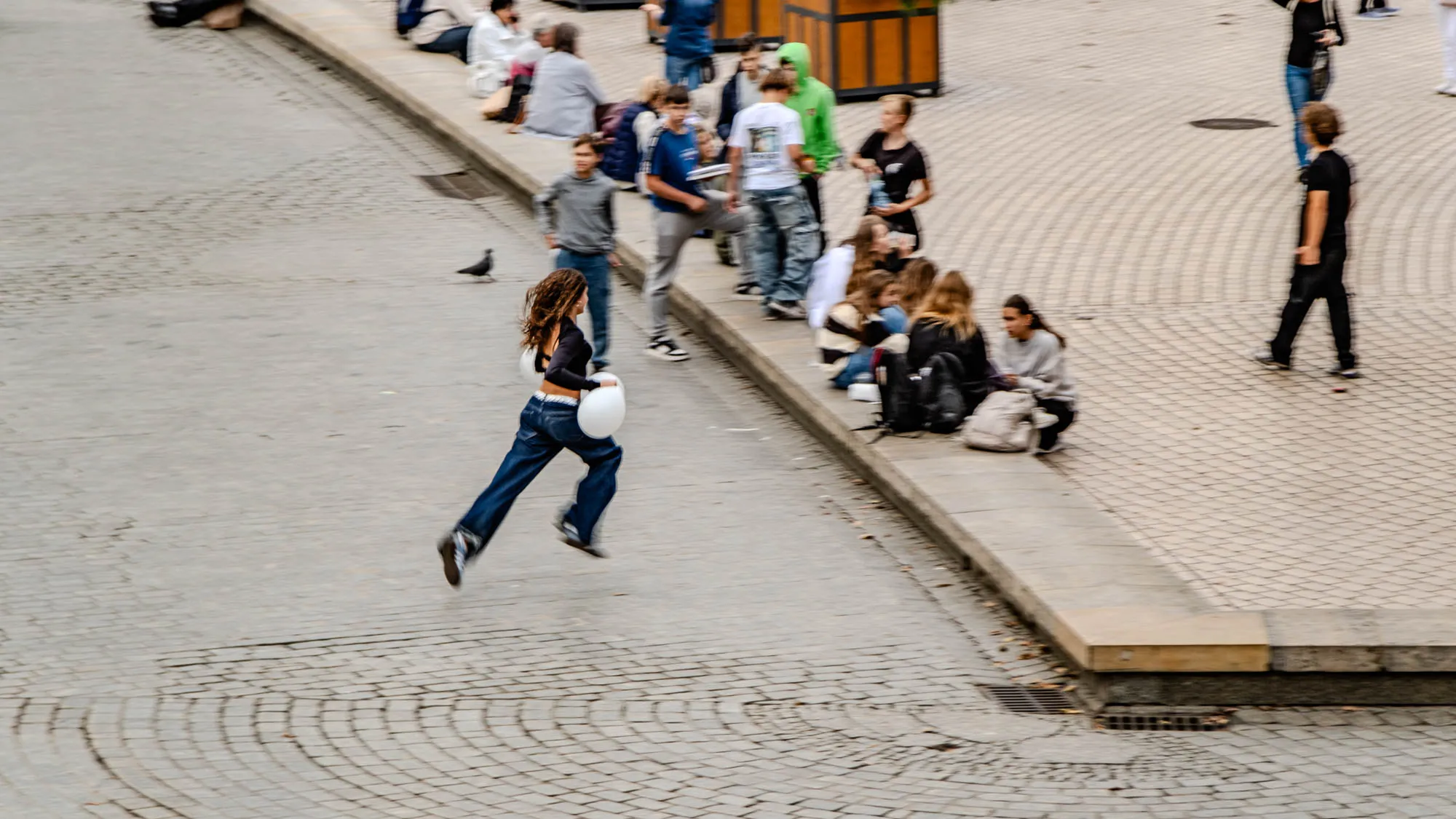 The image shows a cobblestone street with a young woman running down the center of the frame. She is wearing jeans and a black shirt and carrying a white object. There are several people sitting on a curb to the right of her, as well as people walking and standing around the woman in the background. The image is taken from a high angle, looking down at the scene.