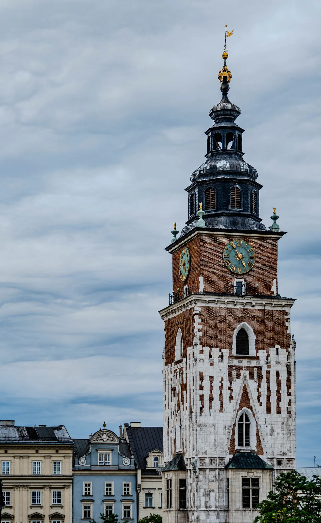 The image shows a tall, slender clock tower made of red brick with white plaster accents. The clock faces are visible on the tower. The tower is topped with a black, shingled, onion-shaped dome and a golden spire. To the left of the tower are three lower buildings with cream and blue-grey facades, their roofs mostly obscured by the tower. The sky above is a hazy blue with wispy, white clouds.