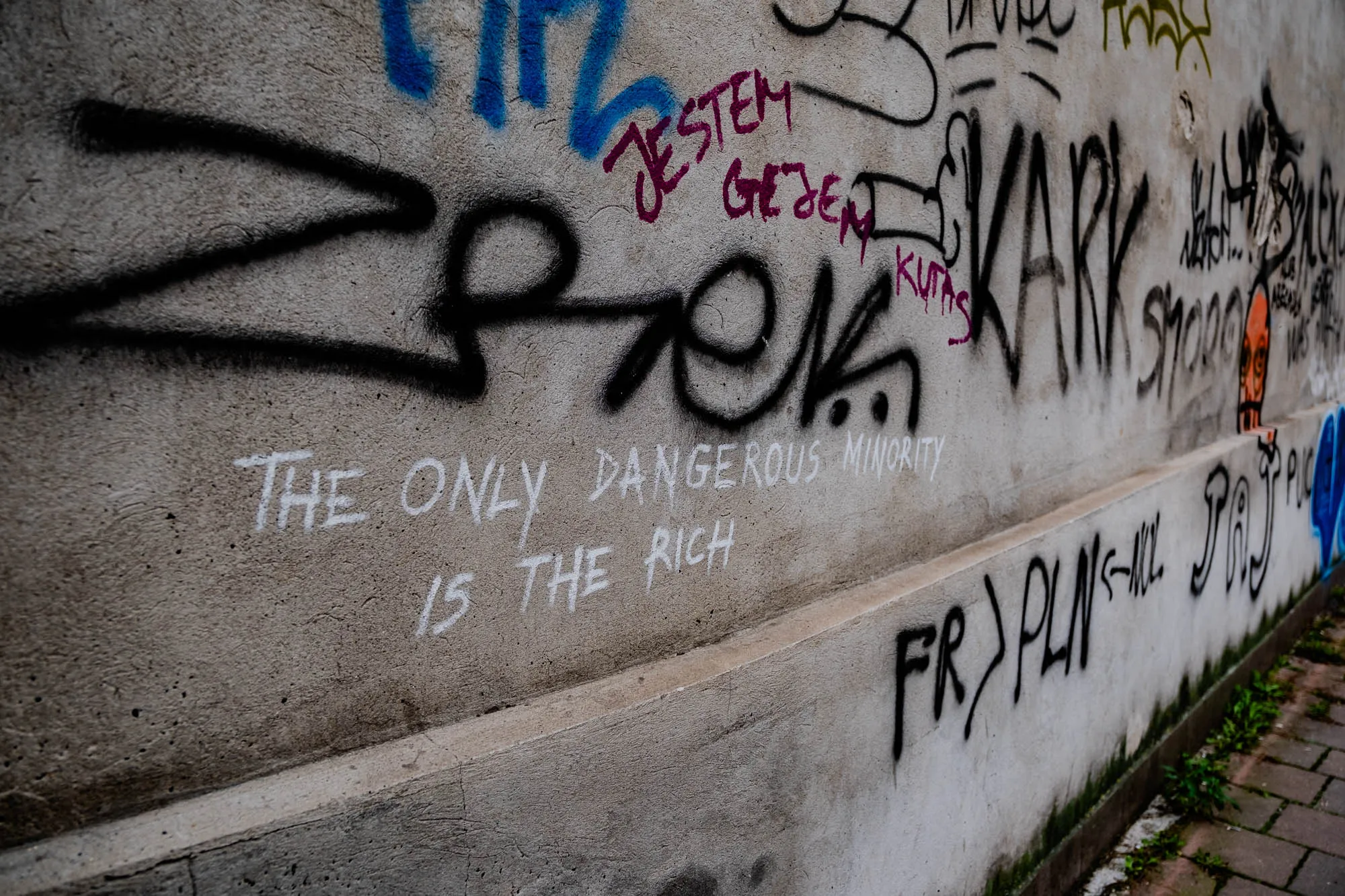 The image is a close-up of a concrete wall covered in graffiti.  The main message in white paint at the bottom center reads "THE ONLY DANGEROUS MINORITY IS THE RICH". Above the message are several other tags and slogans in different colors, including black, blue, pink, green, and yellow. One tag in black reads "KARK" and another in black with a blue outline reads "FR > PLN". There is also some text in pink and black that is mostly illegible, and a partly visible tag in black with a white outline. The wall is rough and textured, and there is a small section of a paved walkway in the bottom right of the image. Some green grass and weeds are growing in the dirt along the edge of the walkway.  The overall impression is that this is a wall in a gritty urban setting. 
