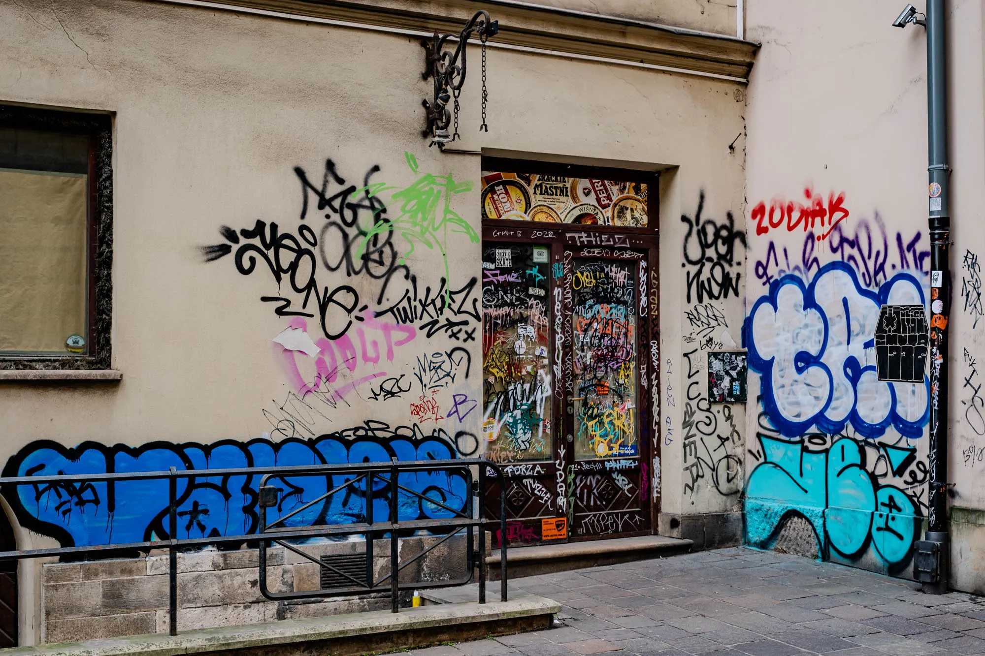 This is a photograph of the side of a building covered in graffiti and posters. There is a door in the middle of the image, which appears to be the entrance to a store or a cafe. The door is brown with glass panels and is covered in stickers and writing. The door is framed by a stone wall with an old wrought iron light fixture above the door, which is hanging from the ceiling. The wall to the left of the door is a pale beige color and is also covered in graffiti and stickers.  The wall to the right of the door is also a pale beige color and covered in brightly colored graffiti. It includes some light blue, dark blue, pink, and black. There is a black metal post sticking out of the wall to the right, on which there is a black sticker and a small, bright yellow and orange sticker.  There is a black metal fence in the foreground, which extends from the wall to the right of the door and down toward the sidewalk in front of the door. The sidewalk in front of the building is paved in gray stone.  In the background, the wall to the left of the door appears to have a window covered in a white, opaque material.  The photo was taken on a cloudy day, as evidenced by the dull lighting.