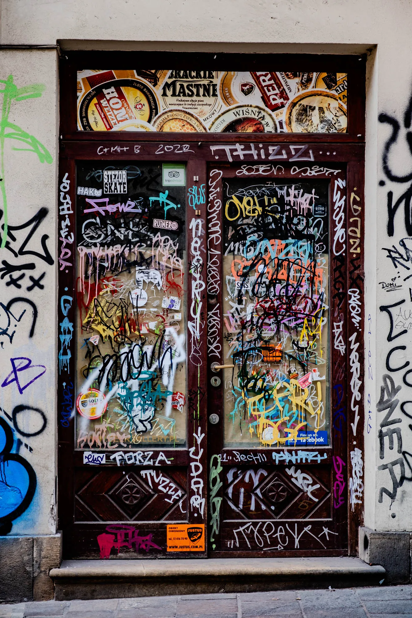 The image shows a wooden double door with glass panels. The door frame is brown with a dark brown frame. The glass panels are covered in graffiti. There is a sticker on the lower left of a glass panel that says "TripAdvisor". The glass panels are covered in graffiti with lots of different colors, mostly blue, pink, green, yellow, orange, and red. The door is set in a white brick wall. There are also white and black graffiti on the brick wall on either side of the door. There is a small step in front of the door. The door is set in a city street with a cobblestone sidewalk in front of it.