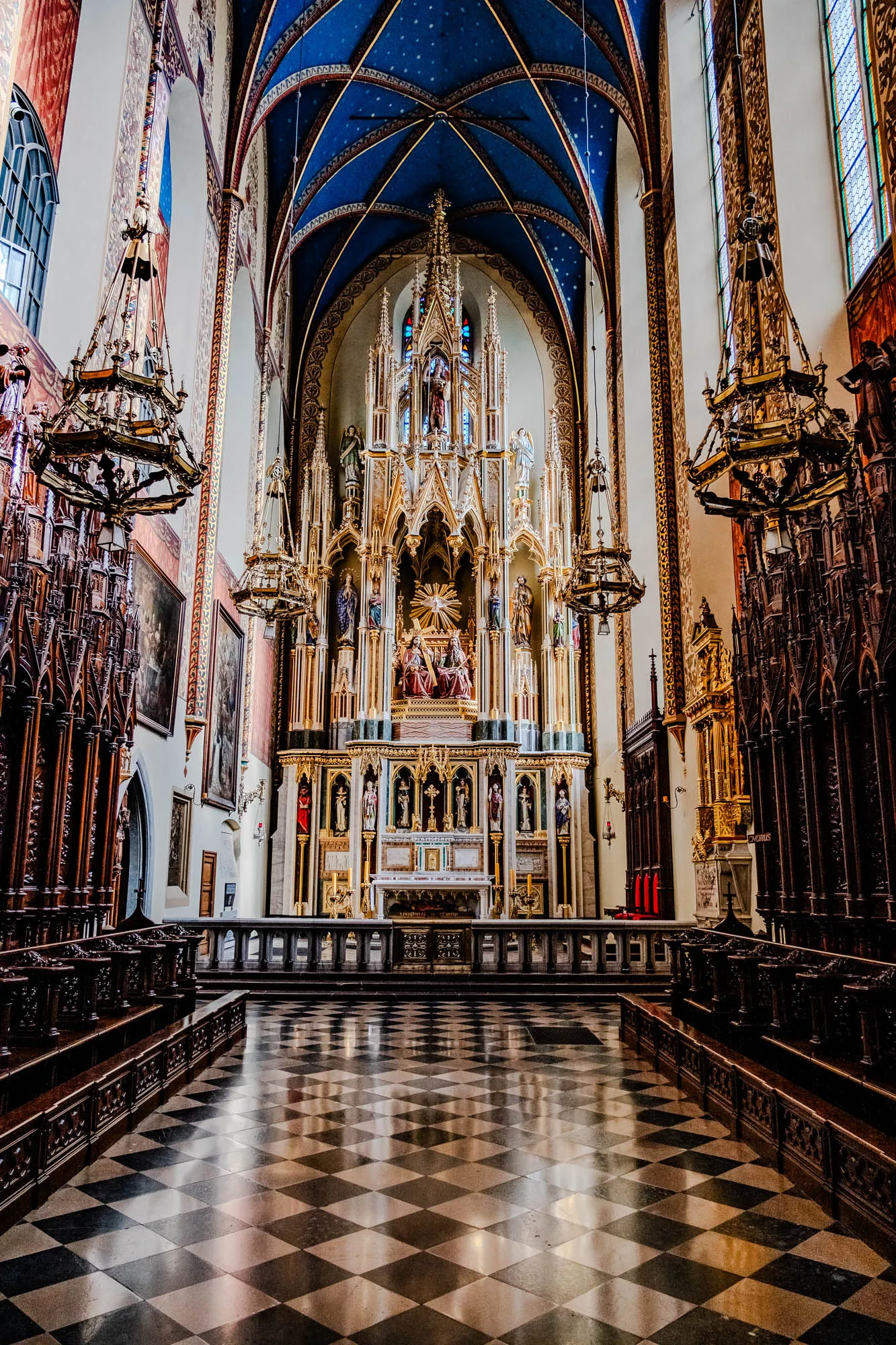 The image shows the interior of a church. It features a grand altar, adorned with intricate carvings and statues.  The altar is made of white marble, with gold accents and carvings. It is flanked by two large, tall candlesticks, each with several yellow candles. Behind the altar, there is a large, ornate wall structure, also made of white marble and gold. It features several statues of various saints and figures, all intricately carved and dressed in detailed robes. The wall structure is also adorned with carvings and paintings. Above the altar, there is a depiction of a dove, symbolizing the Holy Spirit, with rays emanating from it.  Above the dove, there is a large, ornate archway, with more carvings and details. The ceiling of the church is arched and painted in a deep blue, with gold stars. The walls of the church are white, with some painted details and ornate designs. There is a large stained glass window on the right side of the image. The image captures the grandeur and beauty of the church, highlighting its ornate decorations and religious symbolism.