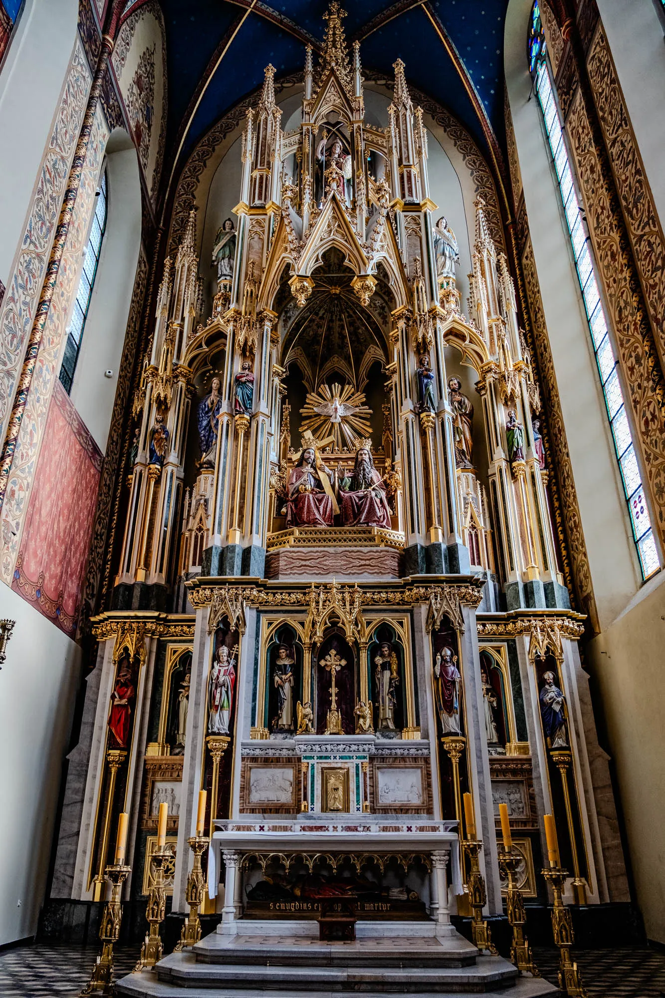 The image shows a large ornate altar in a church. The altar is made of white stone and gold leaf, and is decorated with many statues. The statues are of religious figures, including Jesus, Mary, and the saints. The altar is set against a backdrop of a vaulted ceiling, with a large stained glass window in the background. The walls of the church are white and are painted with floral patterns. There are two large golden candlesticks on either side of the altar, each with three lit candles. The altar is lit from the front by a large light source, casting a warm glow on the scene. At the bottom of the altar, there is a rectangular, stone base with an inscription. The inscription says "SEMPER FIDELIS ET AP MARTYR," which translates to "Always faithful and a martyr."