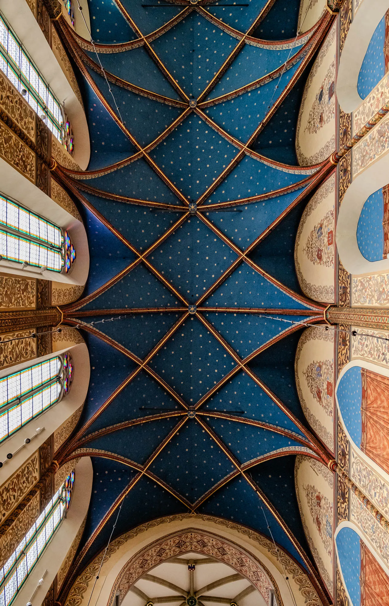 The image shows a vaulted ceiling in a church. It is a blue ceiling with gold trim and small white stars painted on it. There are many arched ribs that create a pattern of squares and triangles. The walls on either side of the ceiling are decorated with painted arches and floral patterns. There are stained glass windows to the left and right of the ceiling. The top of a dome is visible in the bottom of the image. There are several black cords hanging down from the ceiling.  The room is likely inside a church or cathedral.