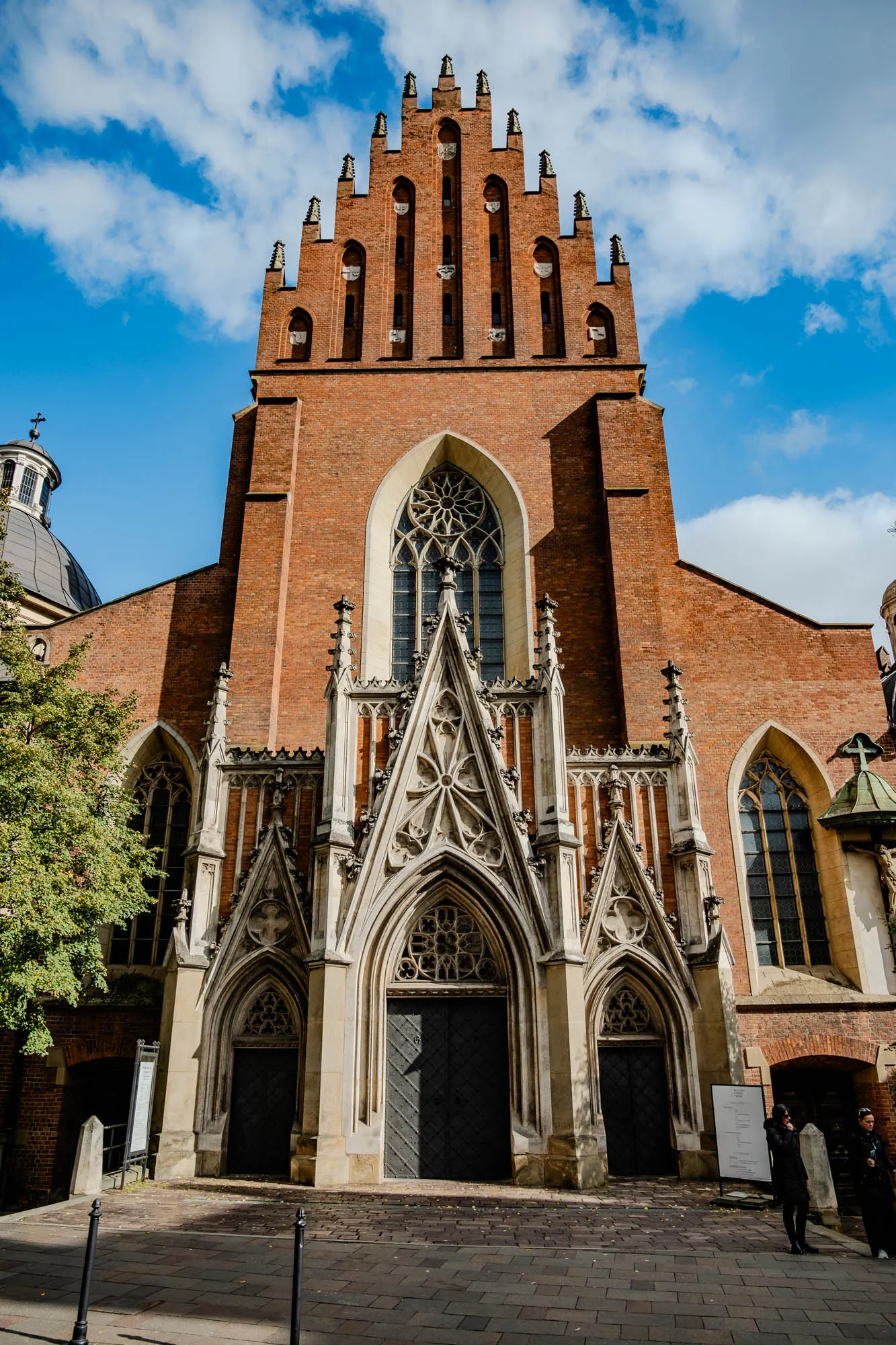 The image shows the front facade of a church. The church is made of red brick and has a tall, pointed archway in the center. The archway is decorated with intricate stonework. Above the archway is a large stained glass window. The church has two smaller doorways on either side of the main archway. The church is set against a bright blue sky with white clouds. The foreground is a cobblestone street with two black metal poles. There are two people standing on the sidewalk to the right of the church, both are wearing dark coats.  To the left of the church is a large tree with green leaves.  The church's dome is visible behind the tree.  The scene is bathed in bright sunlight.  