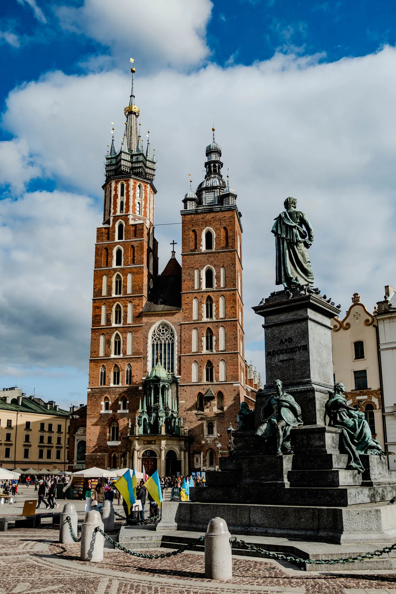 The image shows a large, ornate church with two tall towers topped with spires and decorative gold finials. The church facade is made of red brick with white trim and many arched windows. In front of the church is a large square with a monument in the center. The monument features a statue of a man standing on a pedestal, and it is surrounded by a chain link fence. There are several people standing around the square, and in the background, there is a blue sky with some white clouds. The statue appears to be made of bronze, and it shows a man standing with a cloak.  There are also three other statues around the base of the pedestal, each showing a man and a woman.