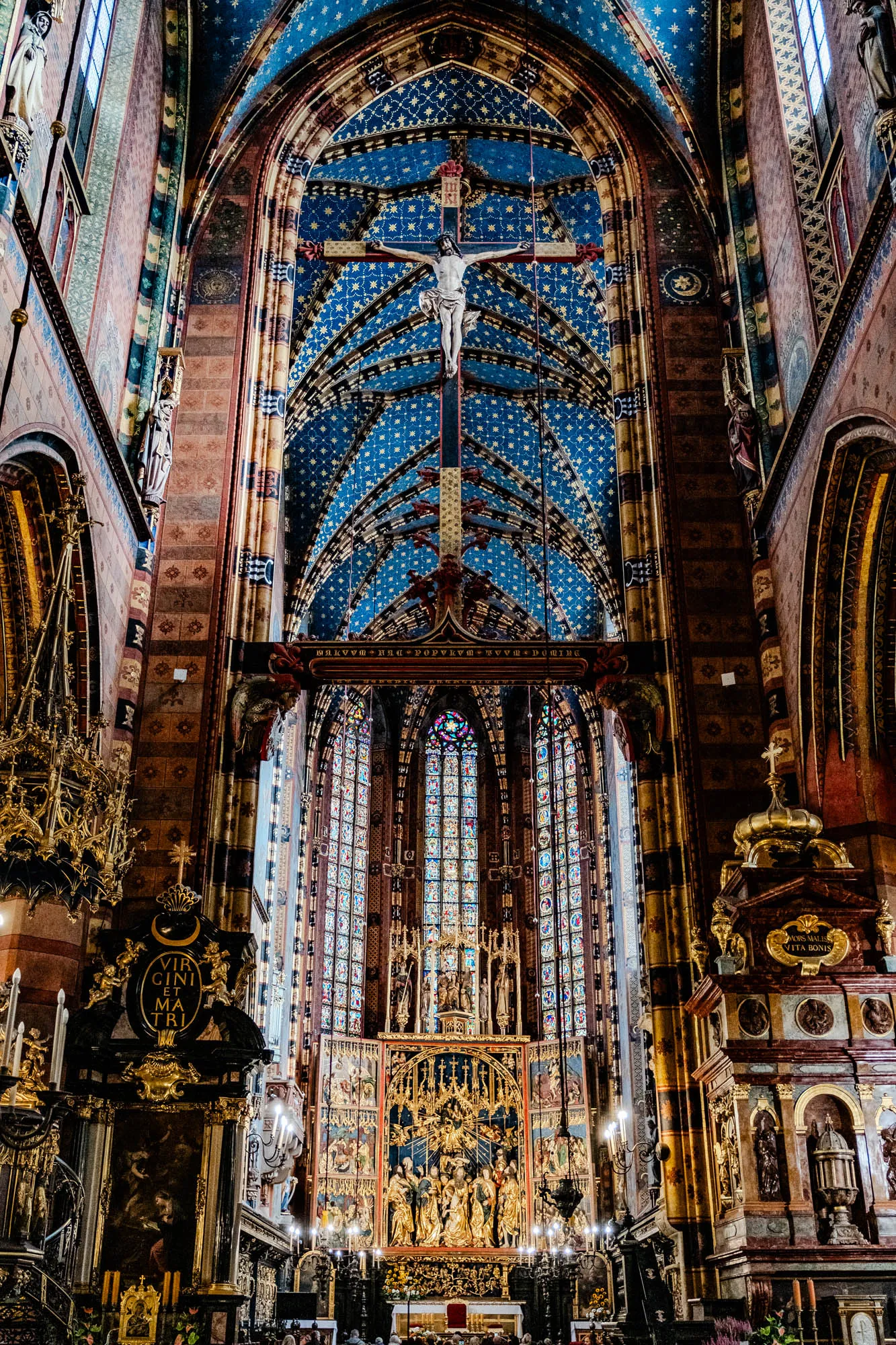 This is a photo of the interior of a cathedral. The ceiling is a deep blue color with gold stars. In the center of the ceiling, a person is nailed to a cross. The walls are made of stone and are decorated with ornate carvings. There are stained glass windows on the right side of the image. There is a large golden altar in the front with a sculpture of people on it. The altar is covered in gold leaf. The altar is in front of the stained glass windows. There are candles lit on the altar. In the front of the image are more people standing and sitting in the pews. There are ornate chandeliers hanging from the ceiling. The walls are decorated with sculptures and paintings. This is a very beautiful and ornate place of worship.