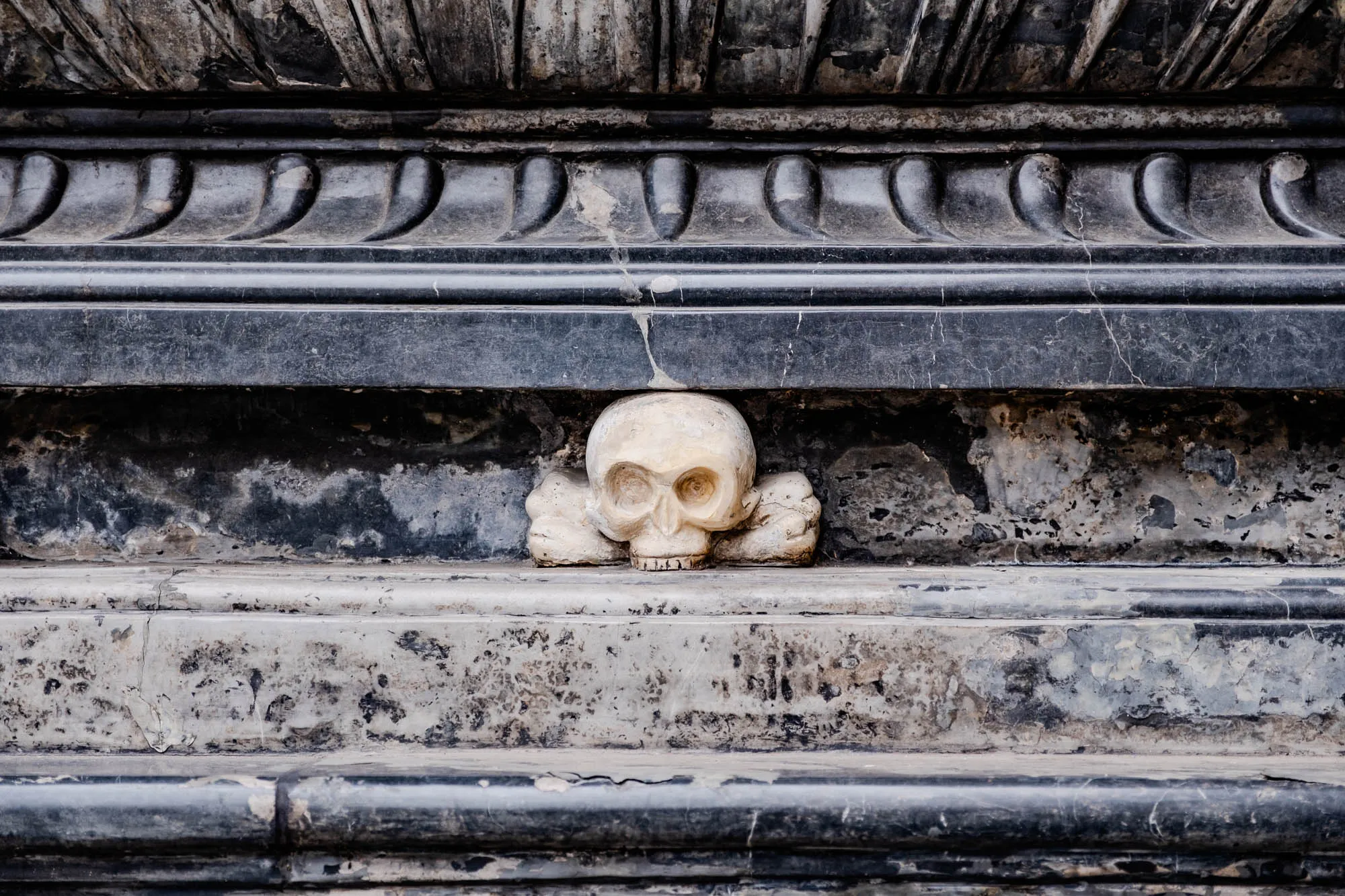 The image shows a close-up of a stone carving depicting a skull and crossbones. The skull is carved in a realistic style, with visible eye sockets, a nose, and teeth. It is made of white stone, which is contrasting with the black marble stone it is carved into. The skull is positioned just above a horizontal stone ledge. Above the skull, there is a decorative stone molding with a series of indentations or curves.  The stone is weathered and worn with age.  The overall tone of the image is dark and mysterious, with the skull serving as a reminder of mortality.