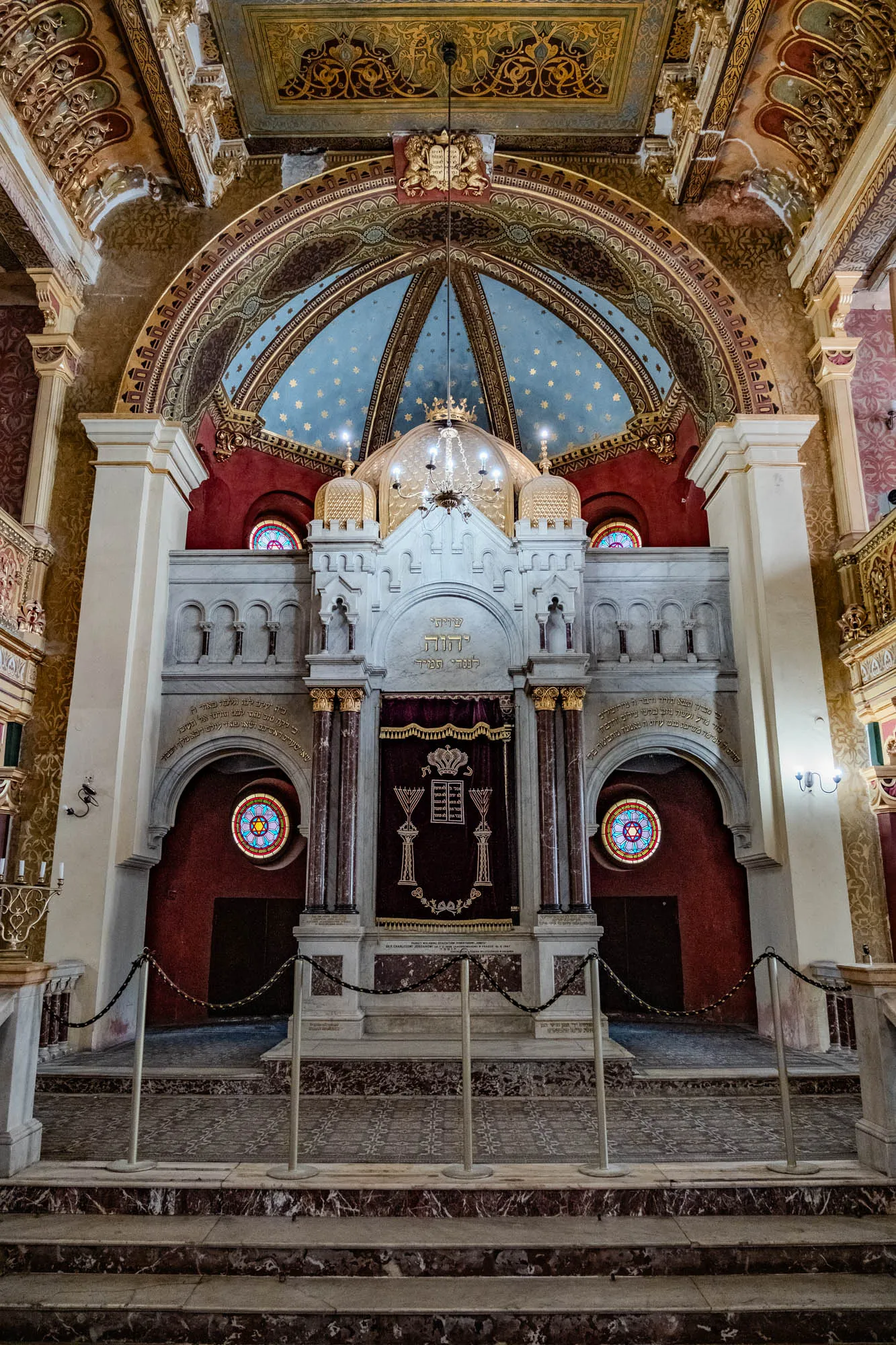 The image shows the interior of a synagogue. The walls are made of white marble and are decorated with intricate carvings. There is a large stained glass window in the back of the synagogue, and a chandelier hangs from the ceiling. The holy ark is in the center of the room, and it is decorated with gold and Hebrew writing. The holy ark is covered with a velvet curtain, and it has a large menorah on it. The holy ark is behind a railing and in front of a set of steps. The interior is dimly lit. In the background you can see a red wall and two more stained glass windows. The ceiling is decorated with gold leaf and intricate designs. The entire room is a beautiful example of architectural detail and design. The holy ark, being the focal point of the image, symbolizes the holiness and sacredness of the synagogue.