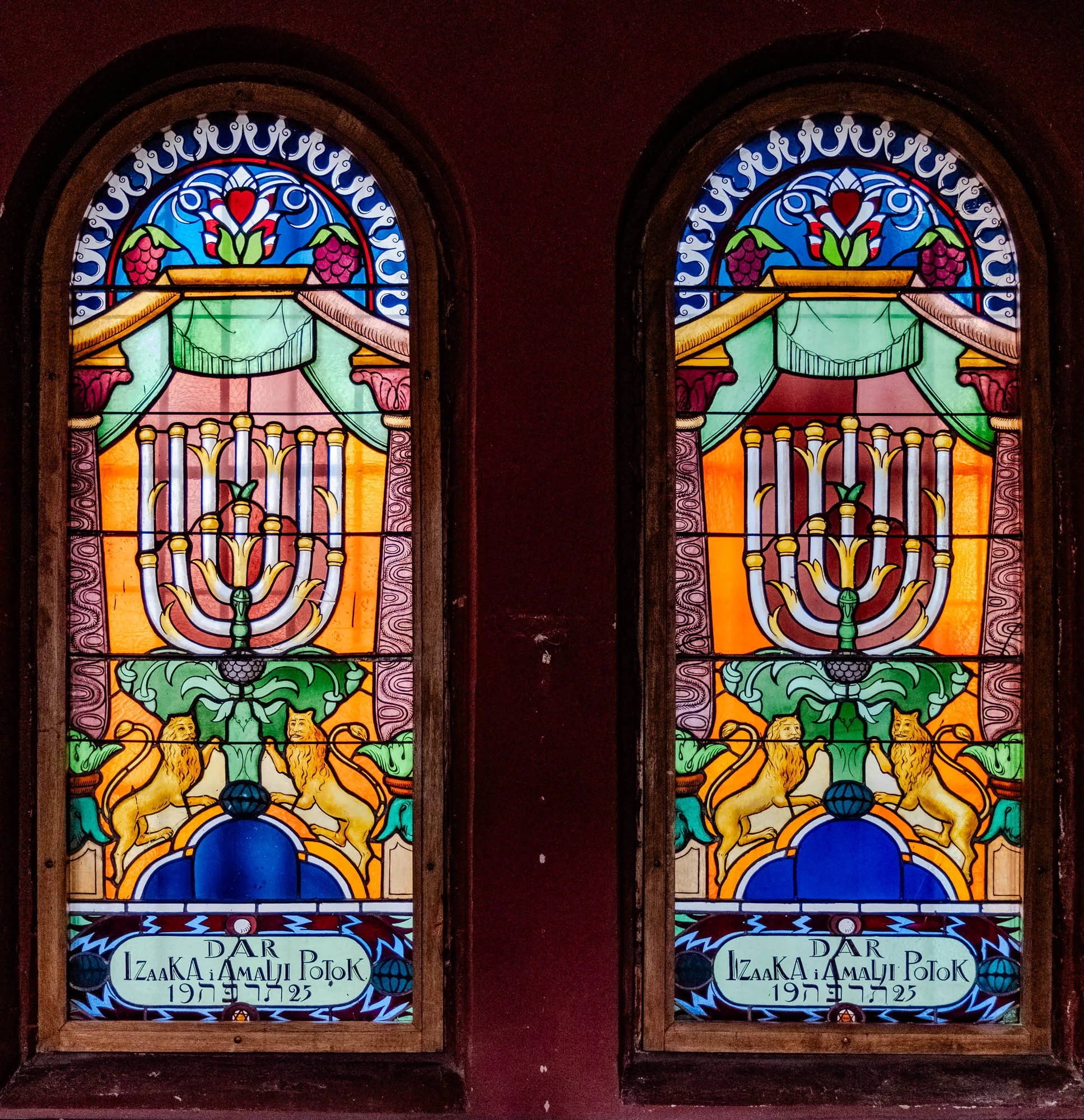 The image shows two stained-glass windows side by side, set into a dark red wall. Each window is an arched shape and is framed by a dark wooden frame. The windows feature a central image of a seven-branched menorah, surrounded by other decorative elements. Above the menorah are two green leaves with a red heart in the center.  The leaves are flanked by two red berries and topped by a blue semi-circle with white swirling details. 

Below the menorah, two golden lions stand on their hind legs. They flank a blue circle with a smaller, blue circle in the center. At the bottom of the windows is a blue inscription with a light green rectangle behind it. The inscription in each window is identical and reads "DAR IZAKA i AMALJI POTOK 1975."  There is a star of David below the inscription, centered within a green circle. The windows are lit from behind, making the colored glass appear vibrant and slightly blurred.