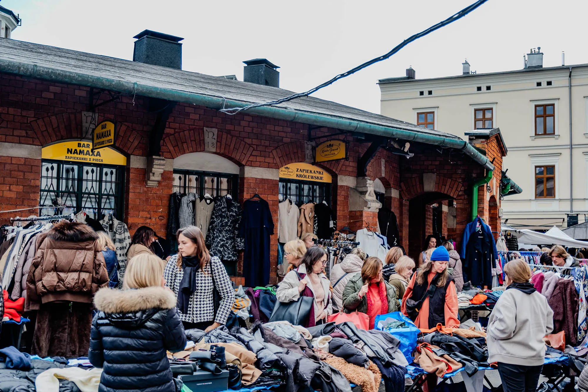 The image shows a street market in front of a brick building. There are multiple tables set up on the sidewalk, filled with used clothing for sale. Many people are browsing the clothes, some are shopping, and a few are talking together. The building is red brick with a green roof. There is a white building in the background with a large, white awning. There are yellow signs above the storefront of the building. One says "Bar Namaxa" and the other says "Zapiekaniki Krolewskie". The image shows a bustling street market in an urban setting.