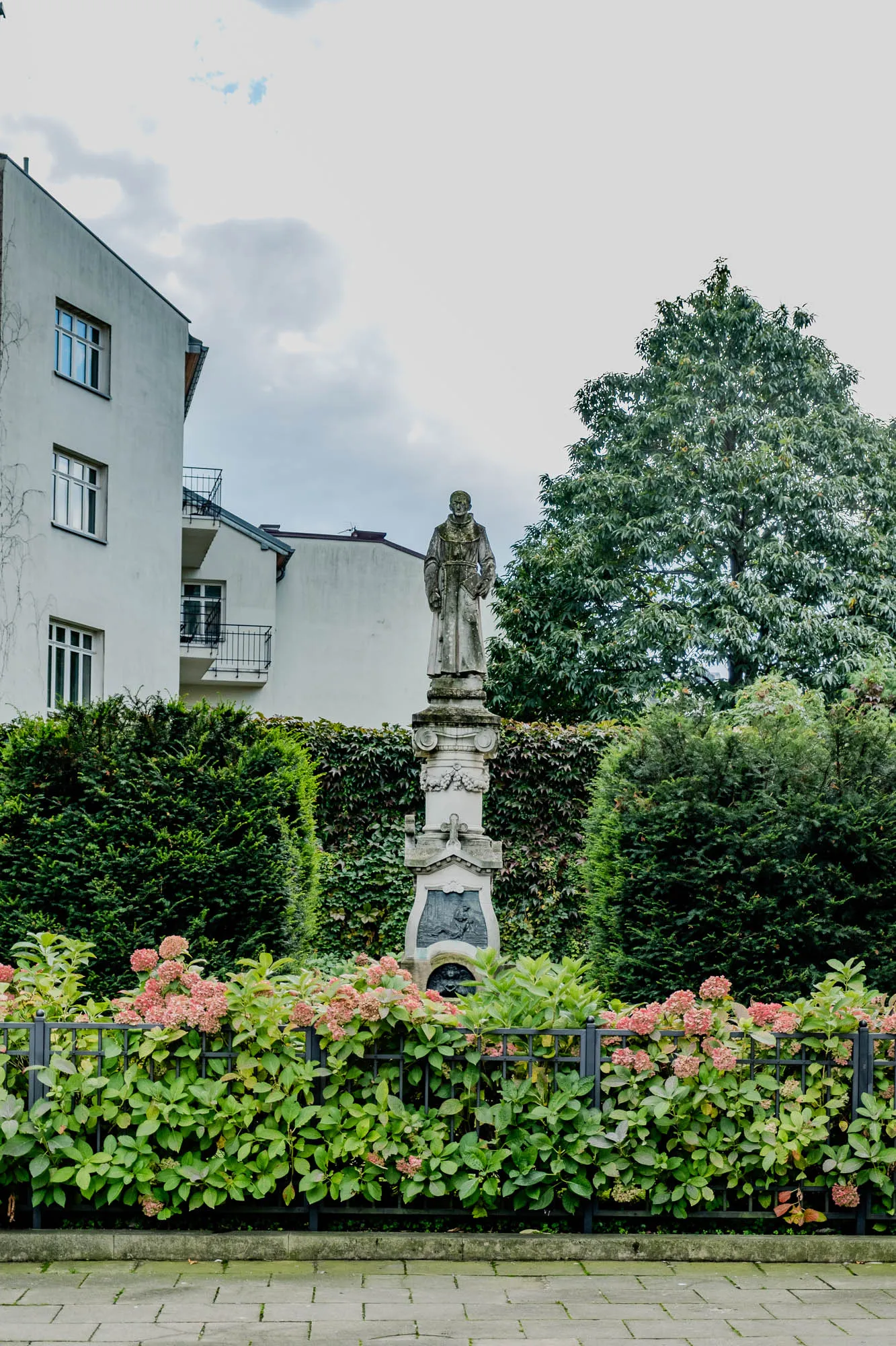 The image shows a stone statue of a person in a robe, standing on a stone pedestal in a park. The statue is located in the middle of the image, with a white building behind it. The building has a balcony and windows. There is a large tree on the right side of the image, and several bushes surrounding the statue. The statue is in front of a wrought iron fence that is lined with flowers, and there is a paved walkway in front of the fence. The sky is overcast with a light grey color. 
