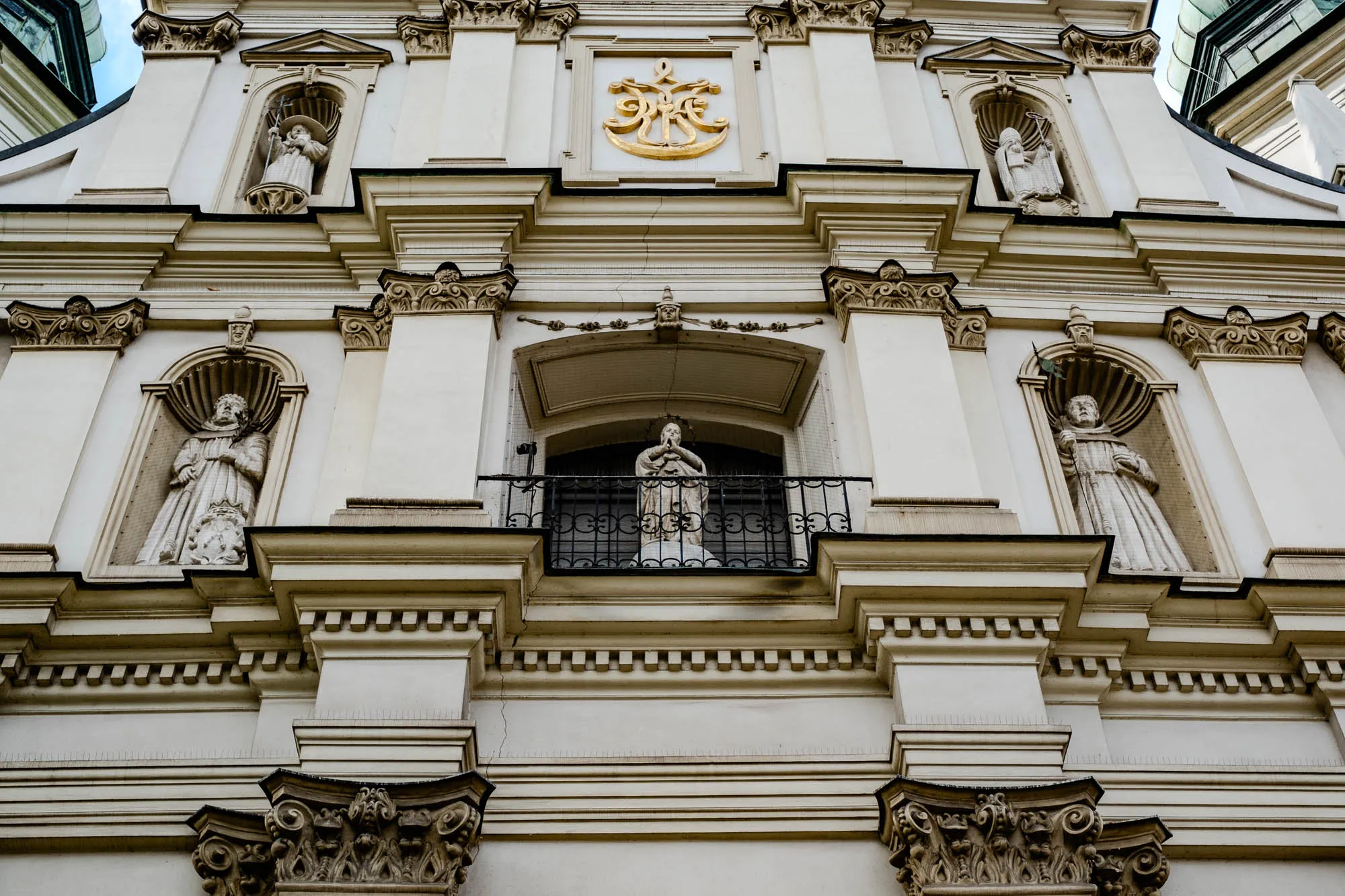 The image shows a close-up of the facade of a building, likely a church or cathedral. The building is constructed of white stone with intricate ornamentation. At the center of the image is a large, ornate gold emblem. Above and below the emblem are niches that house sculptures of religious figures, presumably saints. Each niche is framed by intricate details, including curved arches and ornate mouldings. The figures are carved in white stone and appear to be dressed in flowing robes. The building is decorated with a series of horizontal moldings and cornices that add to its architectural complexity. Below the center niche, a black wrought iron balcony protrudes from the building, with another statue of a saint standing on it. The building has a weathered and aged appearance, adding to its historical significance. The overall impression is one of grandeur and spirituality, reflecting the rich history and architectural heritage of the building.  
